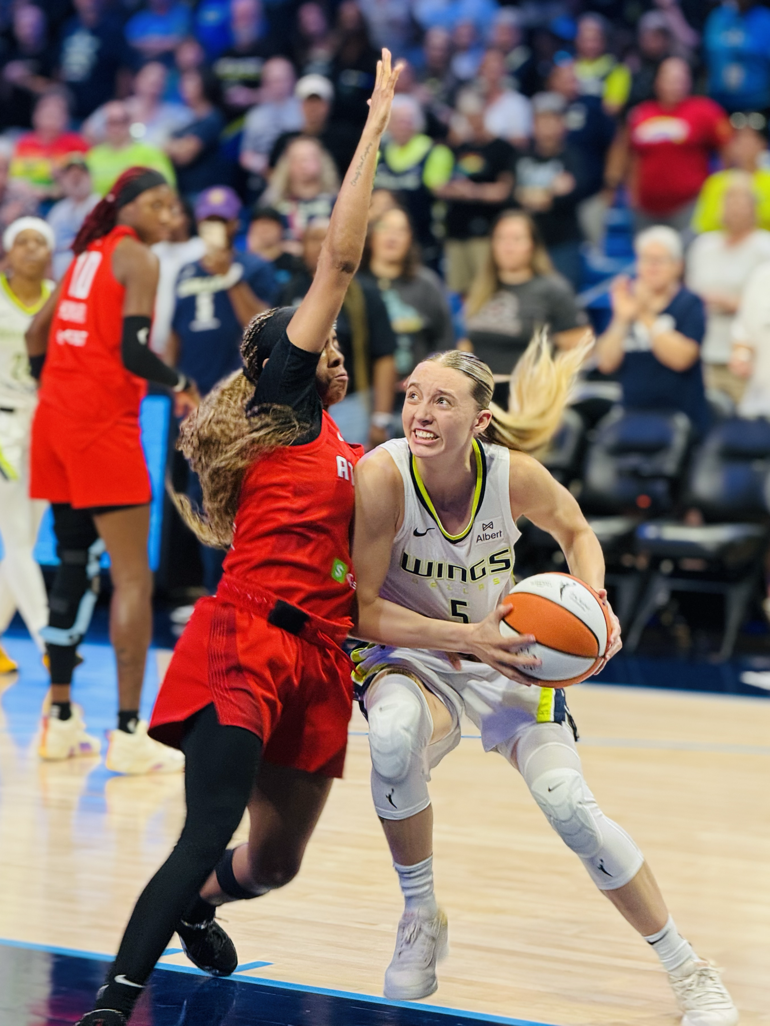Women playing basketball, one in a white jersey with the number 5 and another in a red jersey, with a crowd watching in the background.