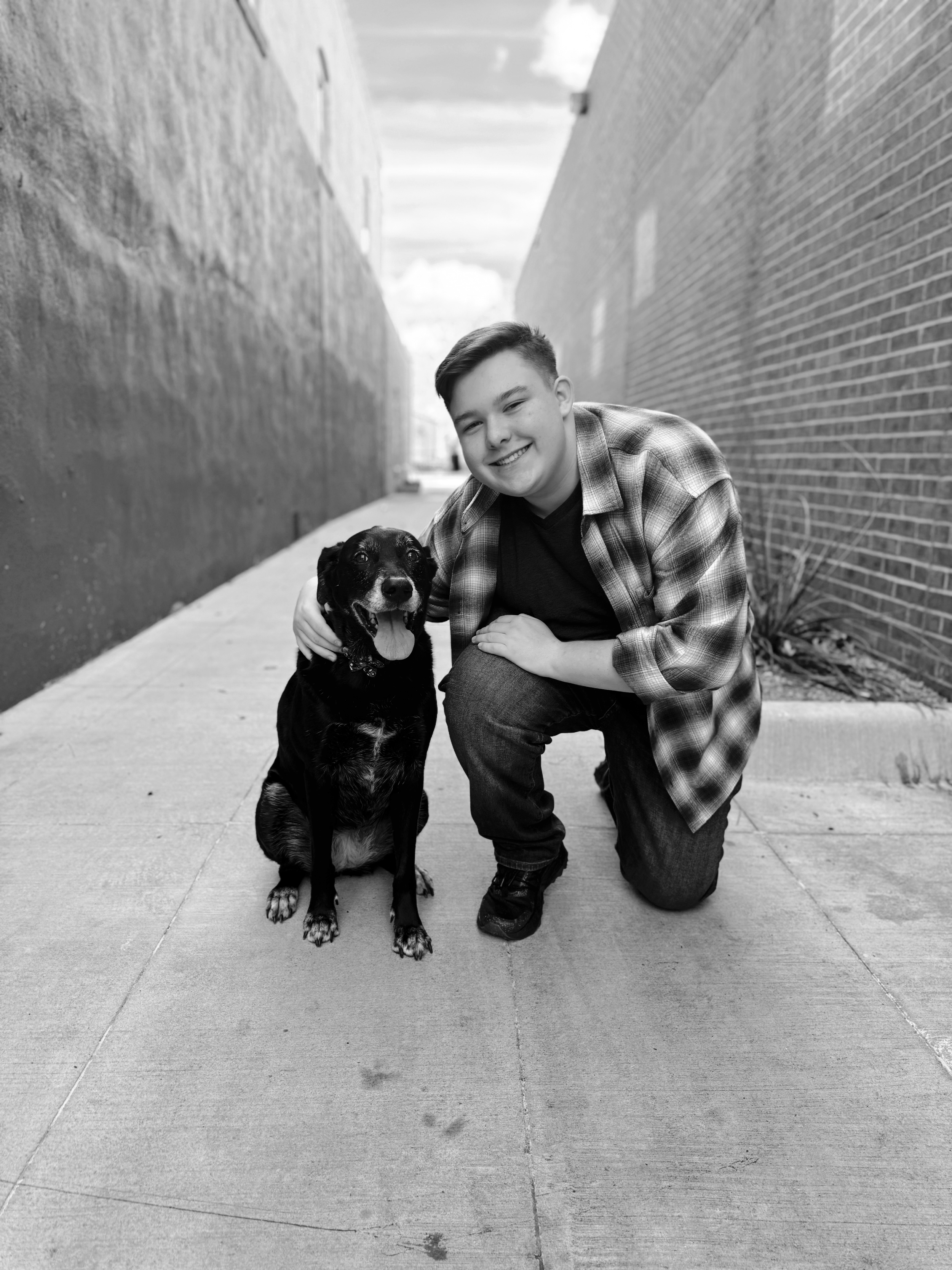A young man kneeling on a sidewalk with a happy dog beside him, in an alleyway with brick walls on either side, in black and white.