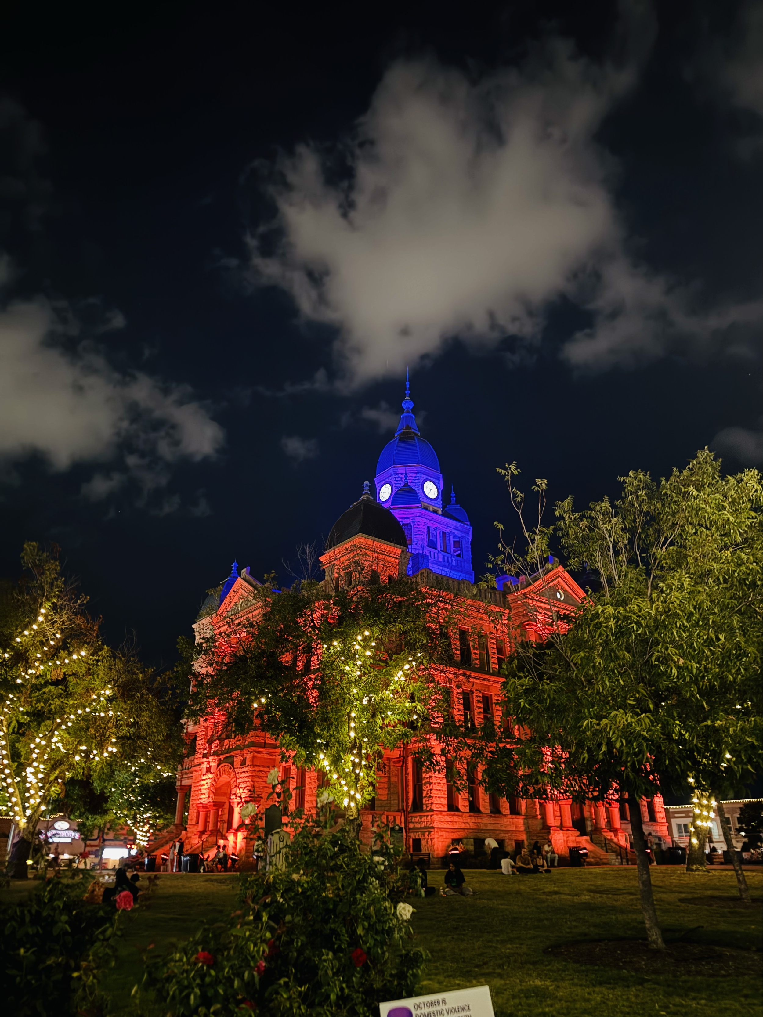 Nighttime view of a historic building illuminated in red, with a clock tower lit in blue at the top, surrounded by trees with string lights, and people sitting on the grass.