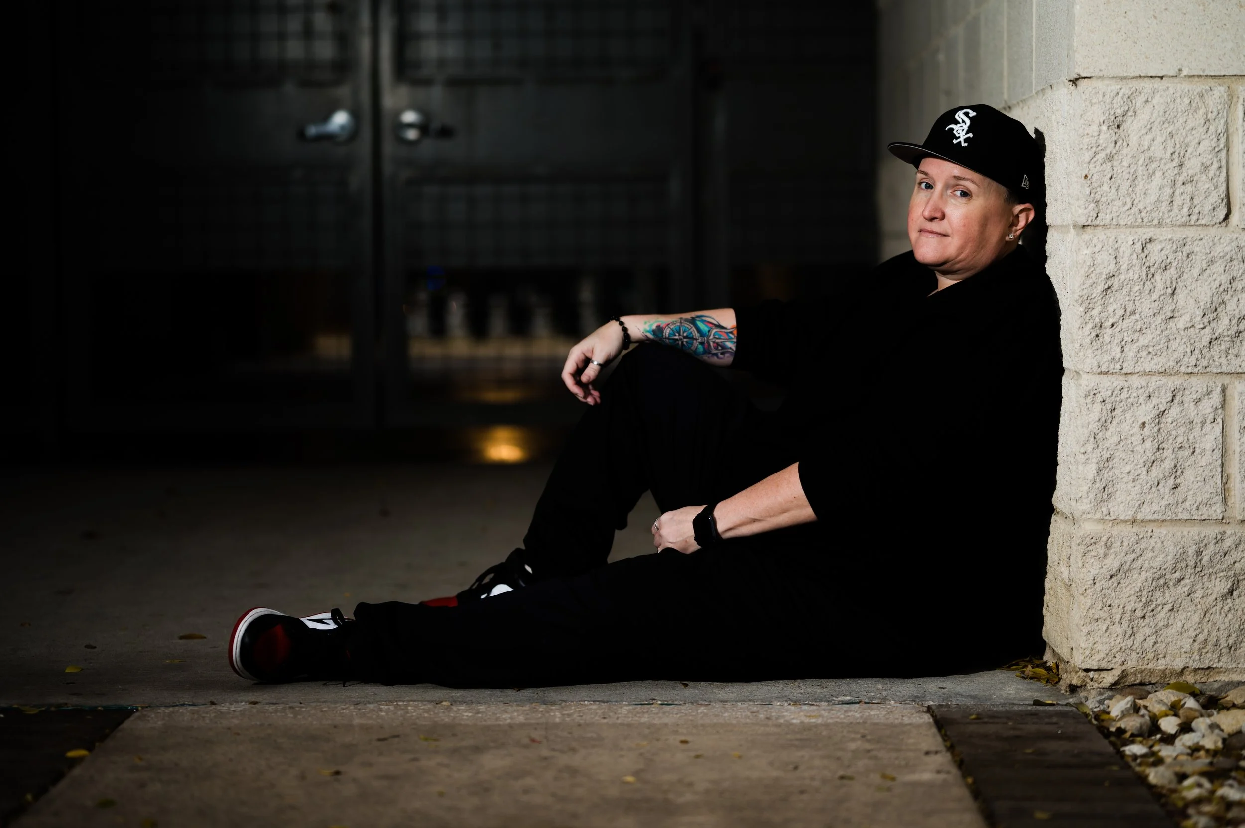 A person sitting on the ground against a stone wall at night, wearing a black hoodie, black pants, sneakers, and a black baseball cap with a white logo, with colorful tattoos on their right arm.