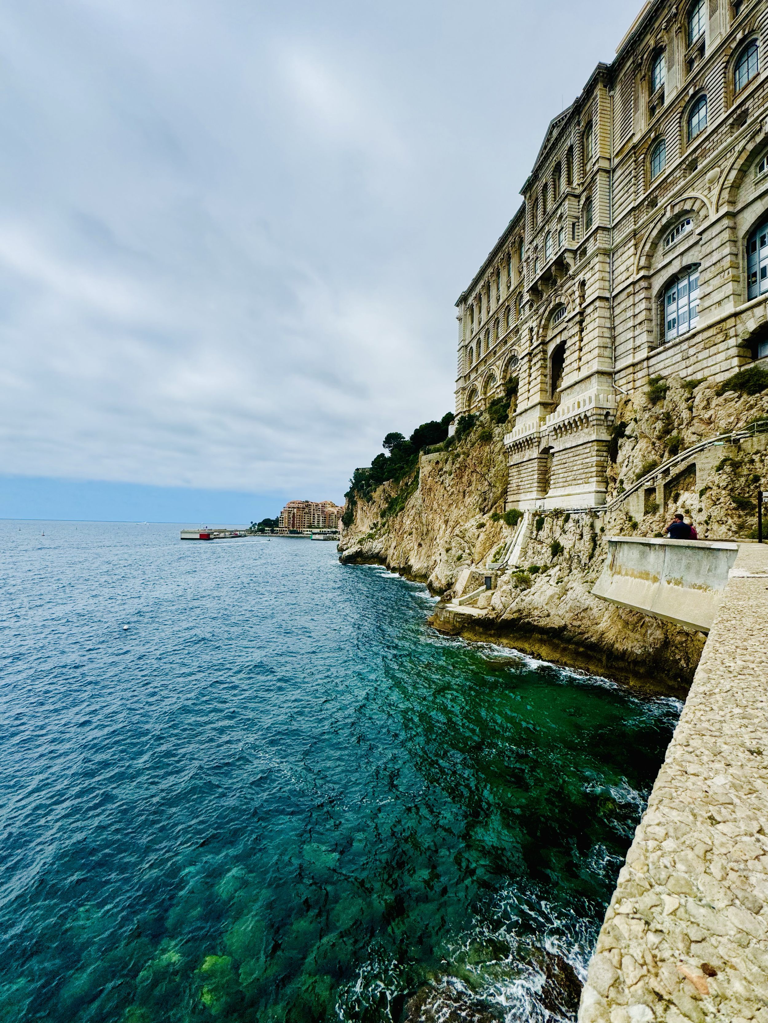 View of a large classic building on a rocky cliff overlooking the ocean, with a pathway and railing along the edge.