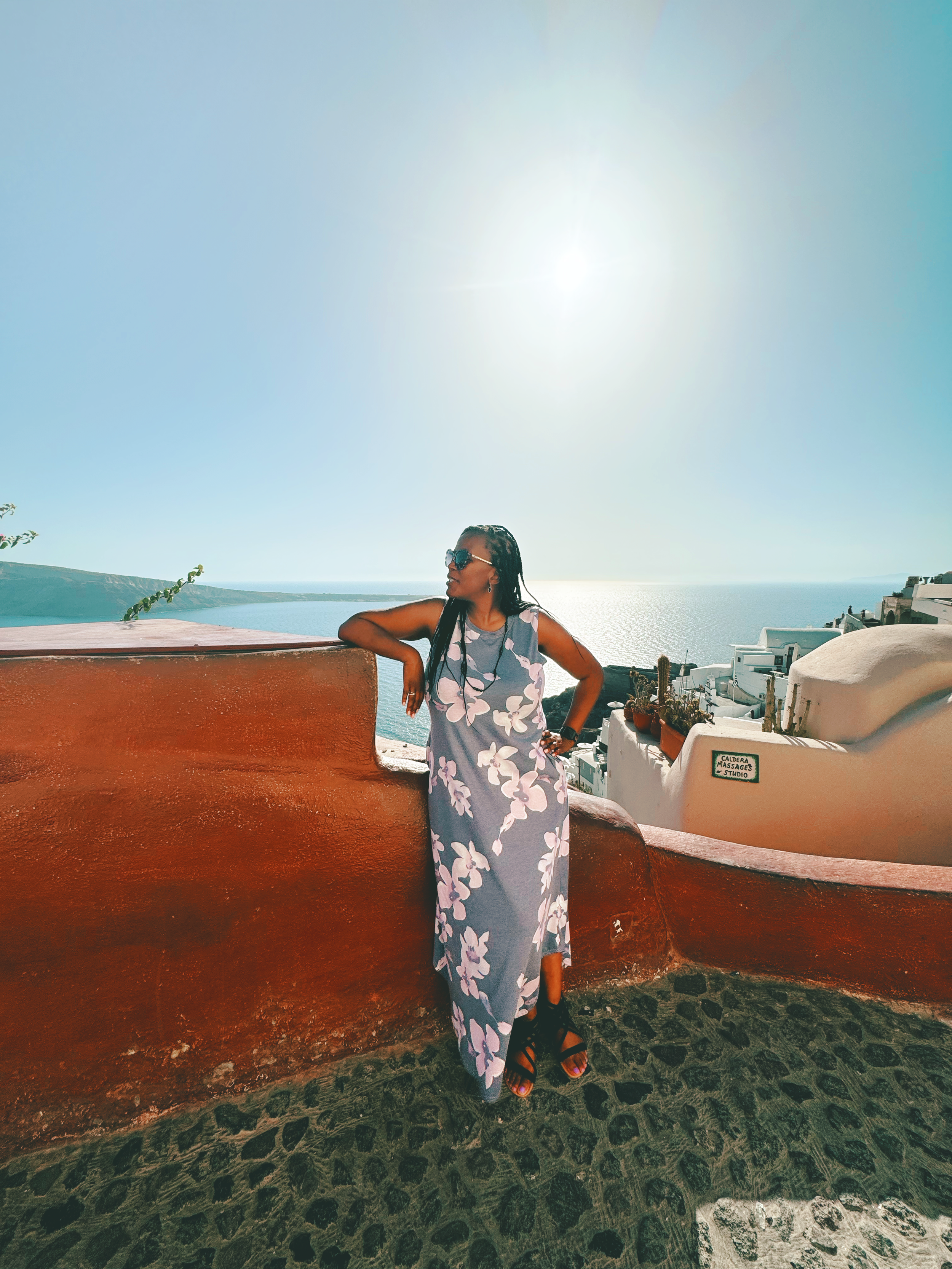 Woman wearing sunglasses and a long floral dress leaning against a red wall on a sunny day with a view of the ocean in the background.