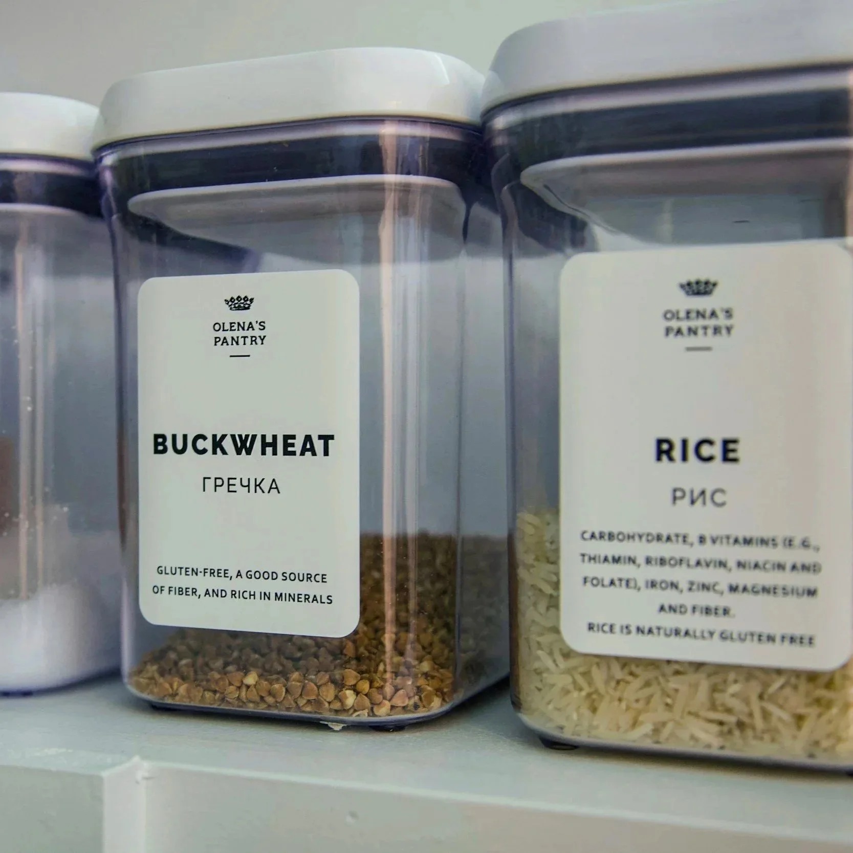 Clear plastic containers with white labels that read 'Olena's Pantry,' containing buckwheat and rice on a shelf.