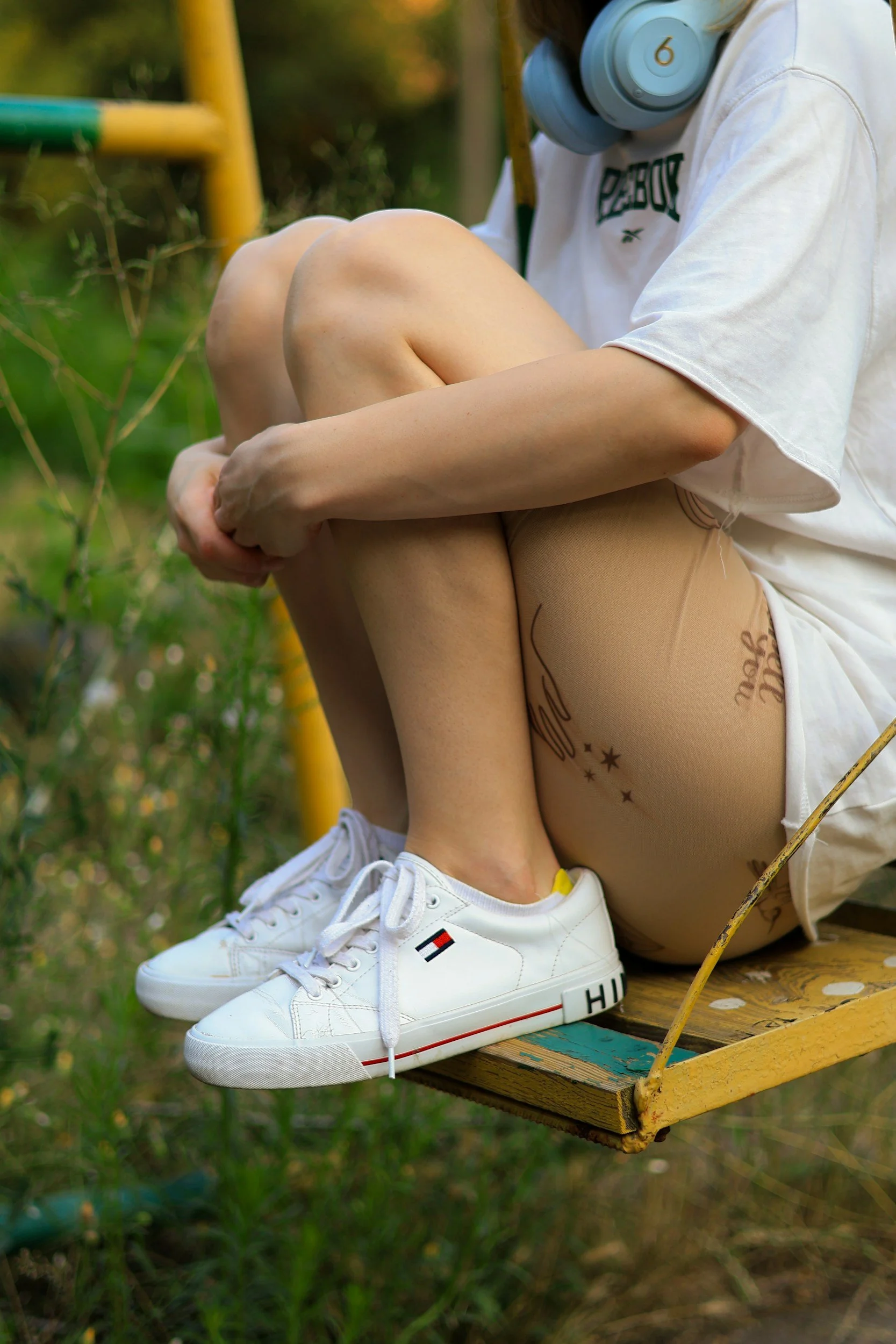 Person sitting on a yellow swing outdoors, wearing beige tights, a white t-shirt, white sneakers, and blue headphones, with tattoos on their leg.