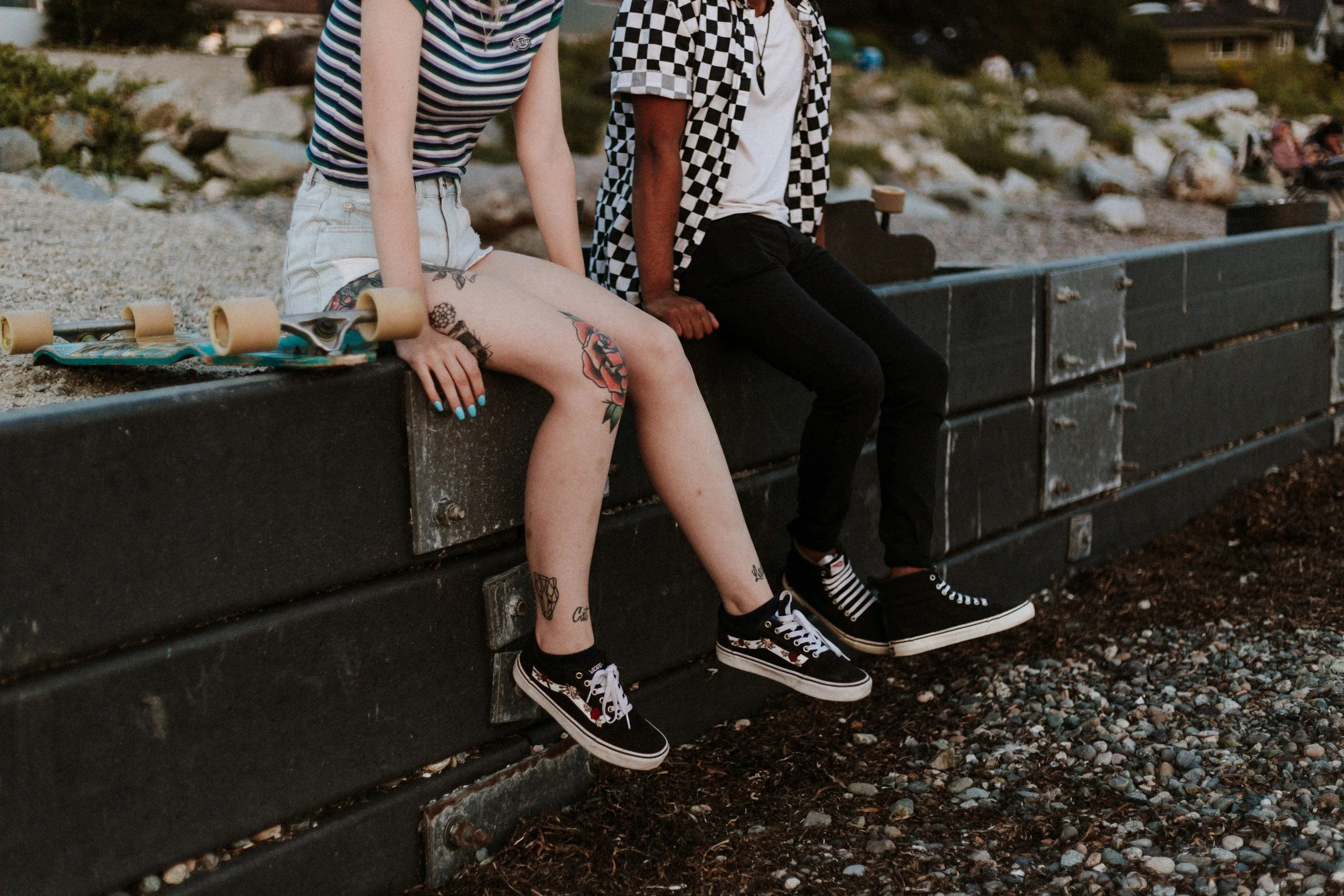 Two people sitting on a concrete ledge at the beach with skateboards; one person has tattoos on their legs and is wearing a striped shirt, and the other person is in a checkered shirt and black pants.