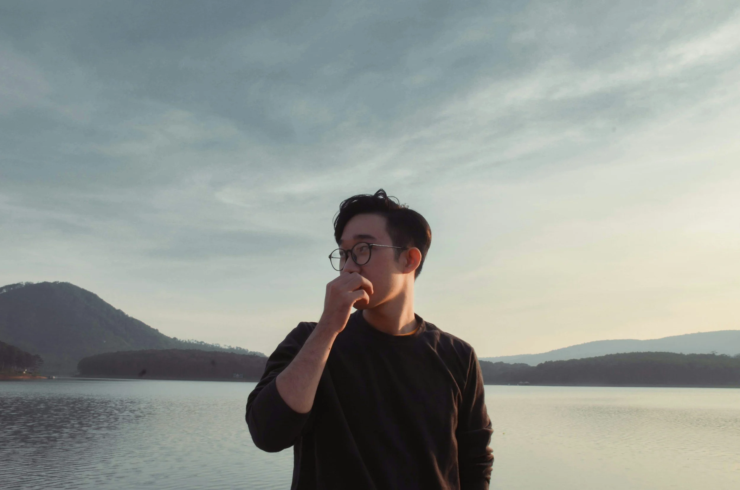 A young man with glasses, wearing a black shirt, standing near a lake with mountains in the background during sunset, with his hand covering his nose or mouth.