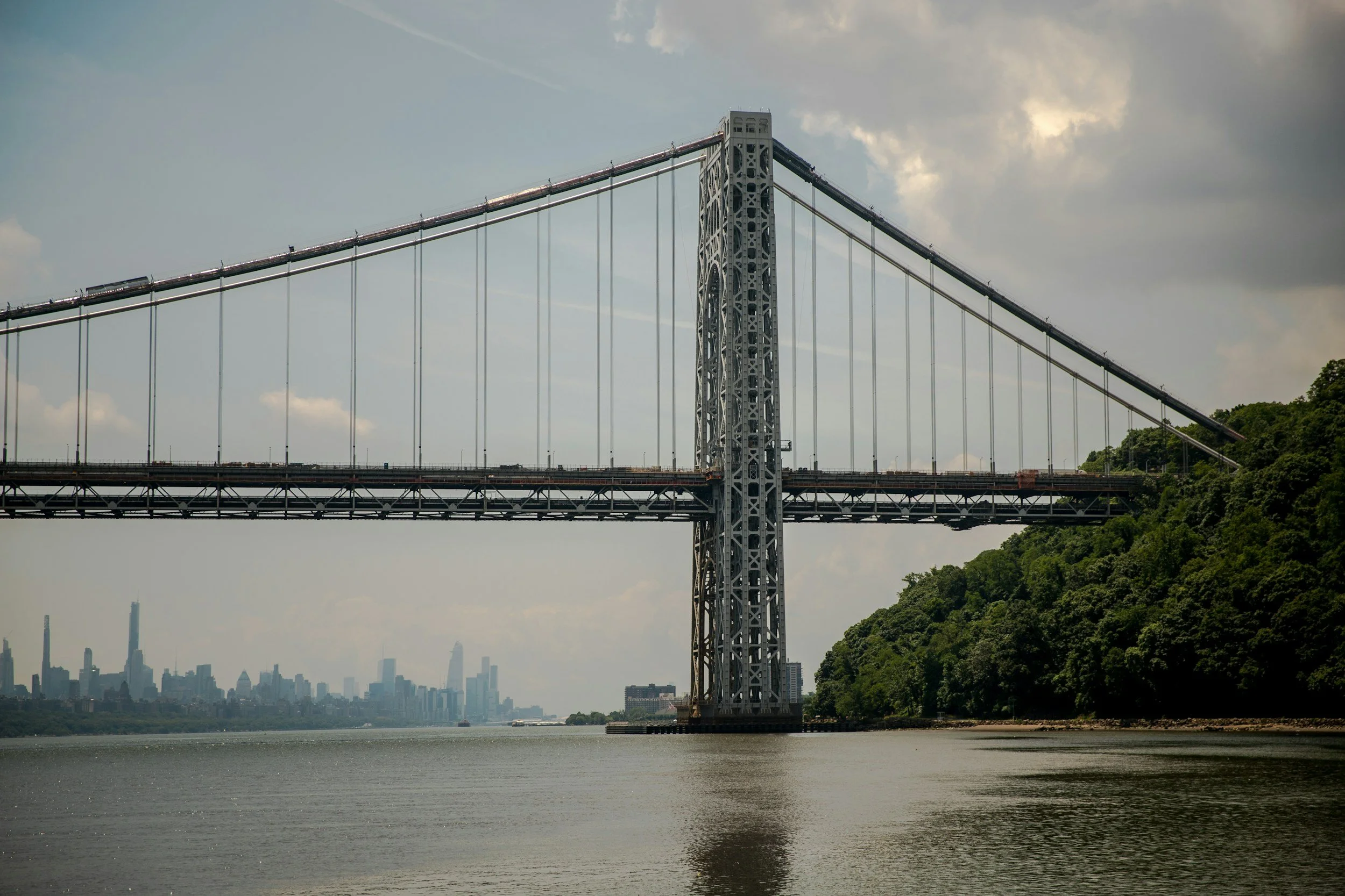 View of the Manhattan Bridge spanning over the water with Manhattan skyline in the distance on a partly cloudy day.