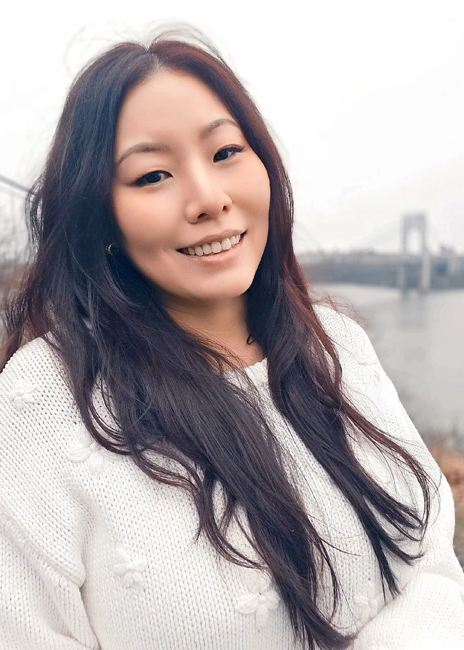 A woman with long dark hair and a white sweater, smiling outdoors near a body of water with a bridge in the background. Therapy for Asian Americans in NYC, Hoboken, Jersey City,  and Bergen County NJ