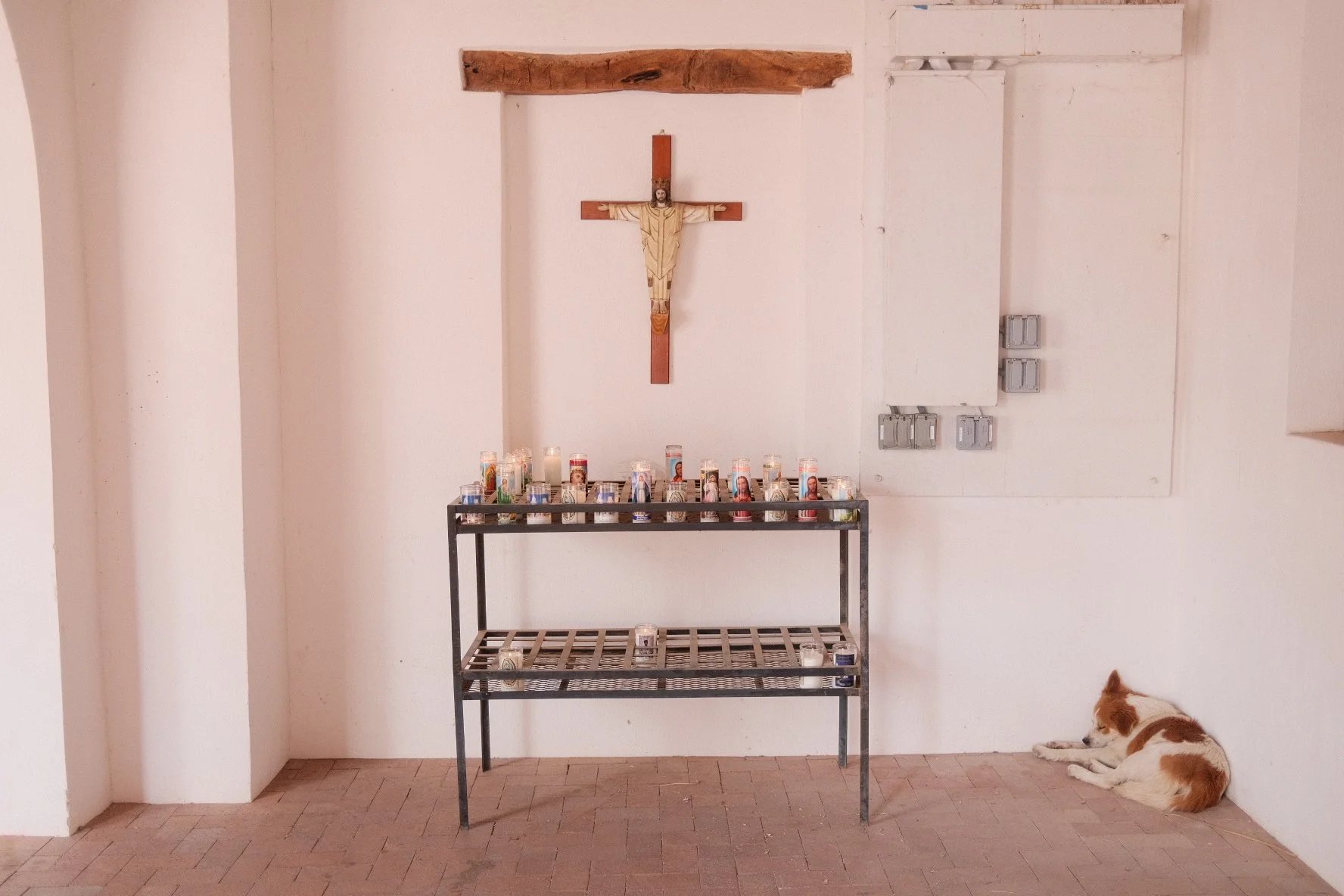 A small altar with a wooden crucifix hanging on a white wall, candles on a black metal shelf below, and a dog lying on the floor to the right.