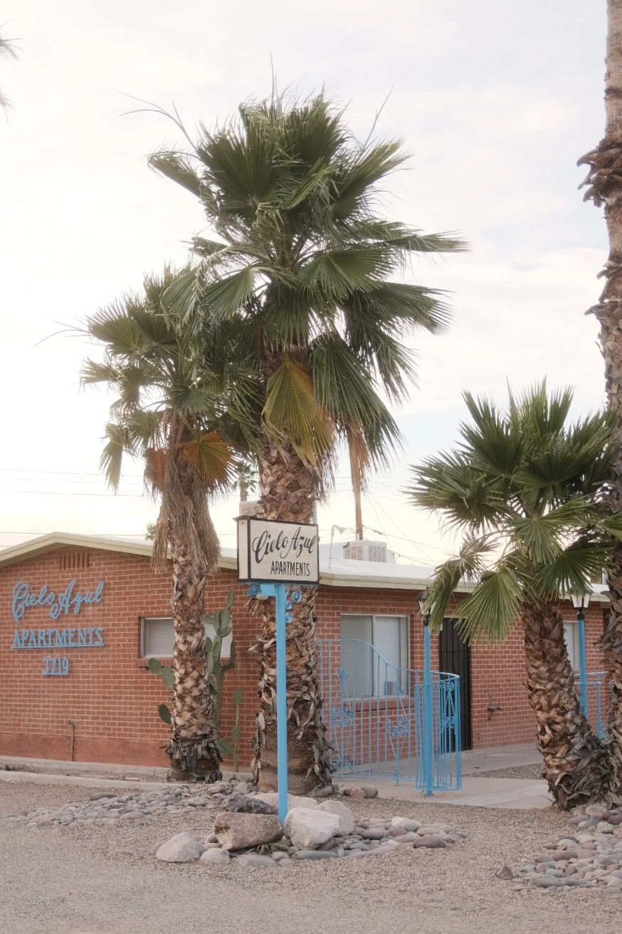 A brick building with blue accents and a sign that reads "Cielo Azul Apartments". The building is surrounded by several tall palm trees and rocks, with a dirt ground.