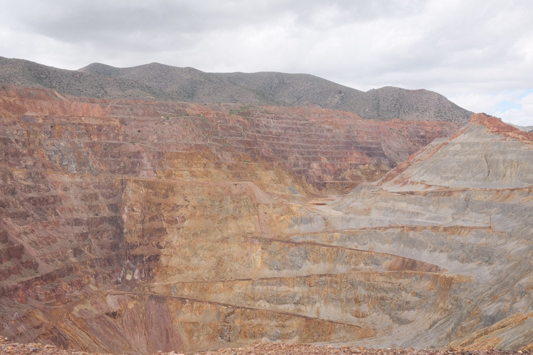 A large open-pit mine with terraced walls of various reddish and brownish minerals, set against mountains and a cloudy sky.