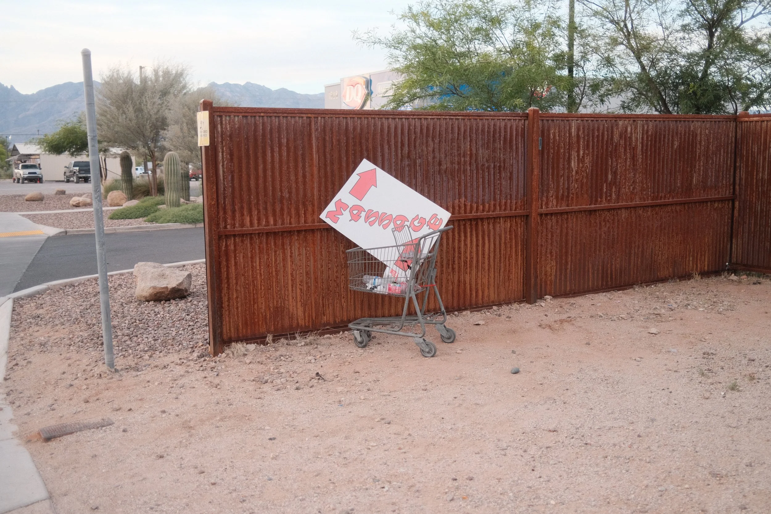 A shopping cart tipped over with a large white sign that has red and pink text and an arrow, positioned in front of a tall, rusty, brown metal fence.