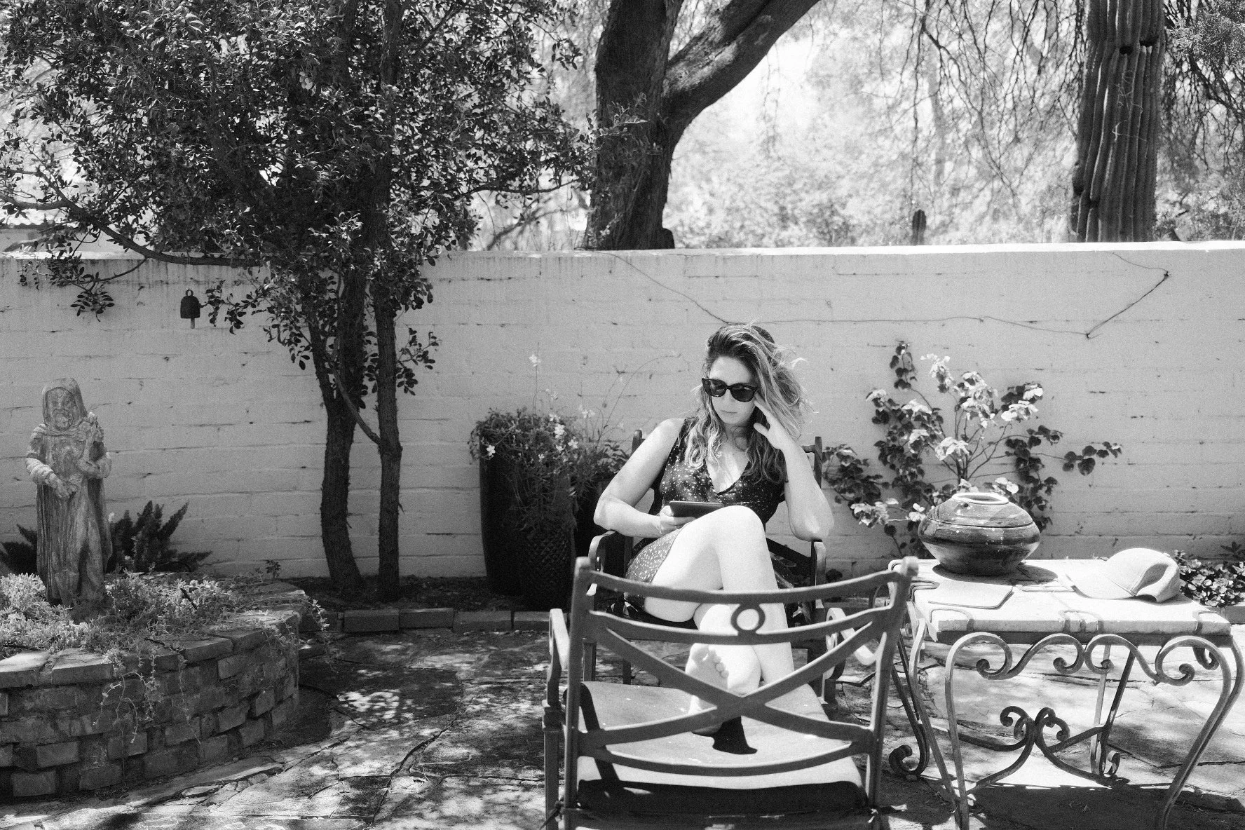 A woman with wavy hair wearing sunglasses and a dress sitting outdoors on a patio, reading or using a device, surrounded by plants, garden statues, and outdoor furniture in a backyard setting.