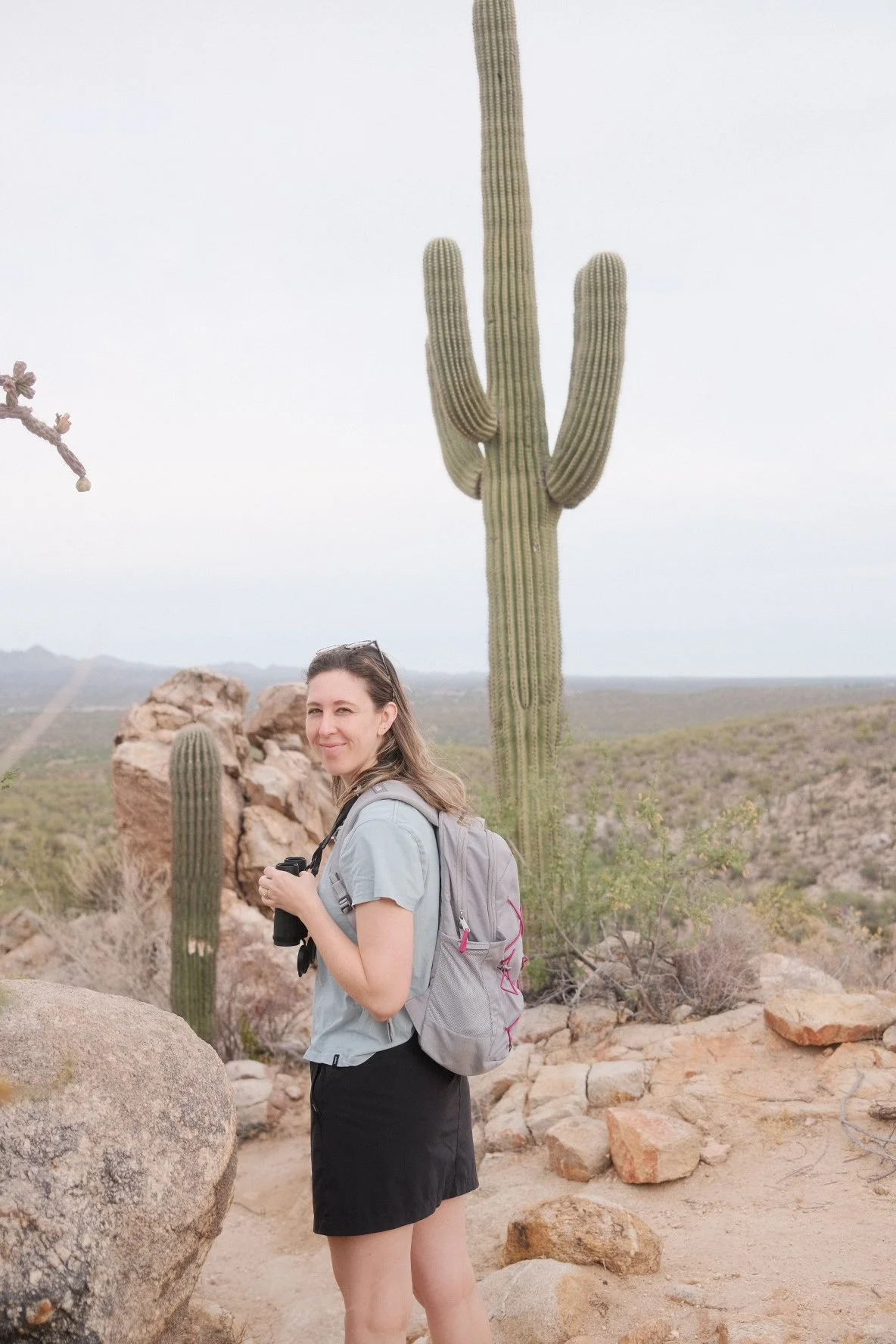 A woman with a backpack standing in a desert landscape with rocks, cacti, and mountains in the background.