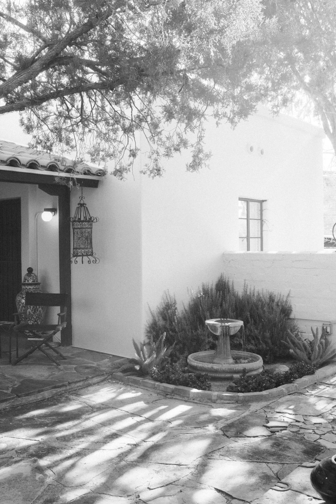 Black and white photograph of a courtyard with a stone patio, potted plants, a small fountain, and a white stucco wall with a window. A large tree with overhanging branches partly shade the area.