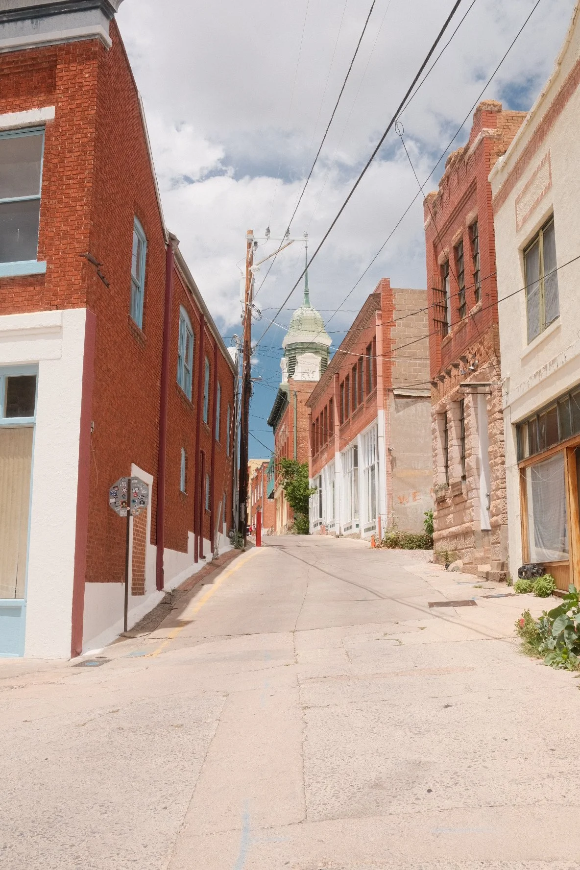 A deserted urban street with brick and concrete buildings on either side, overhead power lines, and a church steeple in the background under a partly cloudy sky.