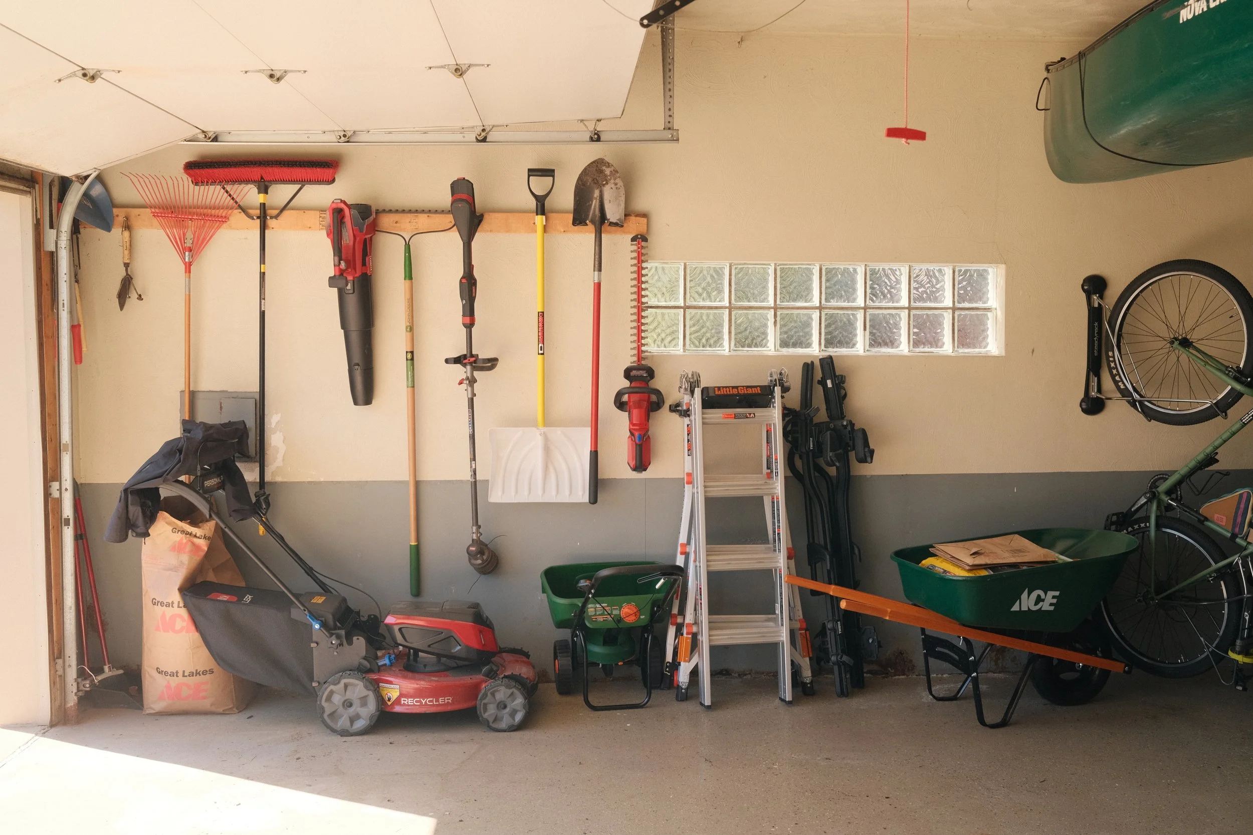 Garage with various yard tools and equipment hanging on the wall, including rakes, shovels, a leaf blower, and a ladder. There is a lawnmower, a bicycle, and a trash bin.