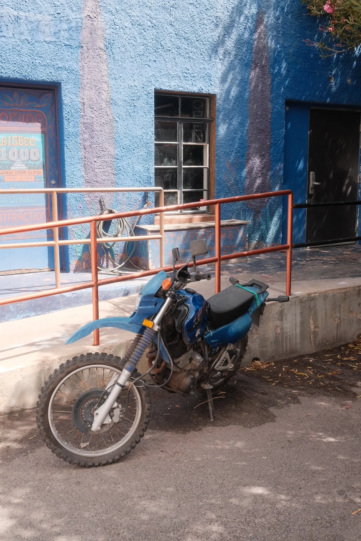 Blue off-road motorcycle parked in front of a blue building with a window and a door. The motorcycle is missing its front wheel and fender, and a green strap is tied around its seat.
