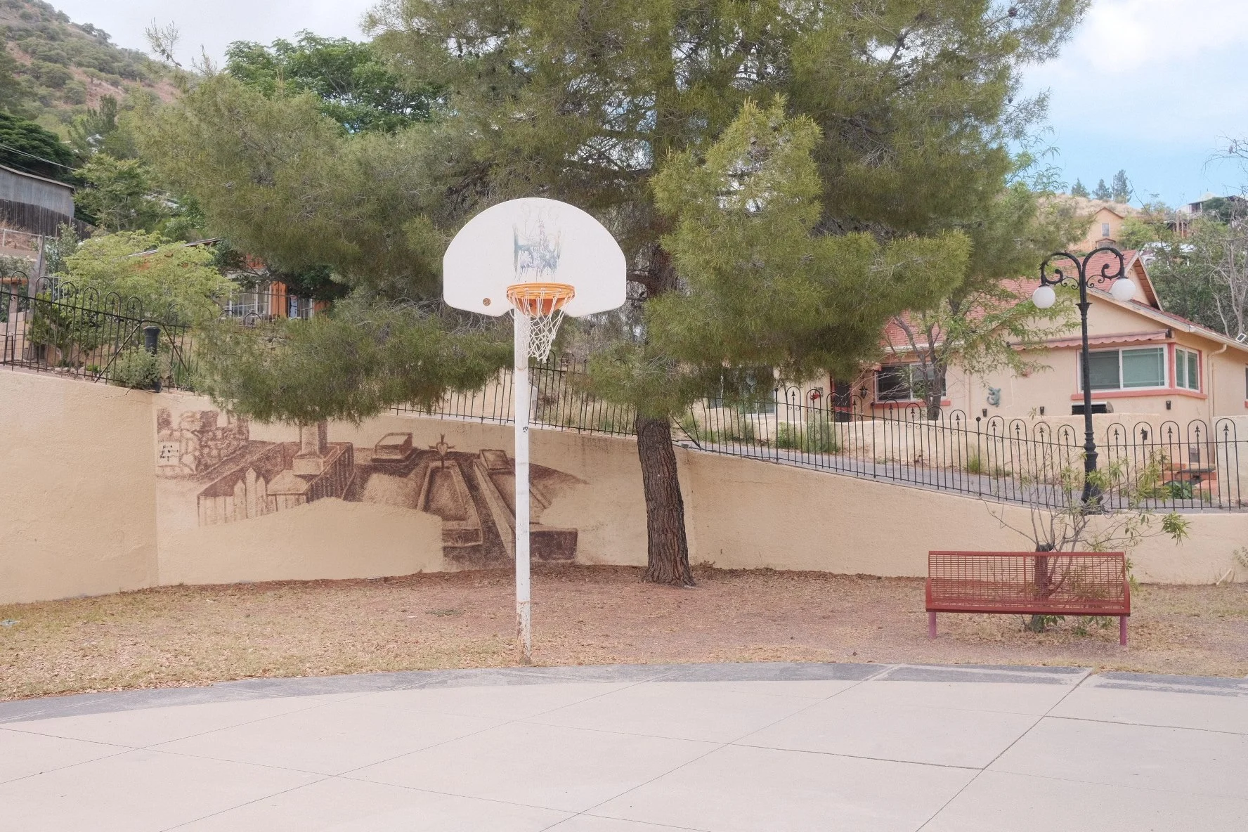 A basketball court with a basketball hoop, bench, tree, lamp posts, and residential houses in the background. There is a mural on the wall depicting a staircase and a fountain.