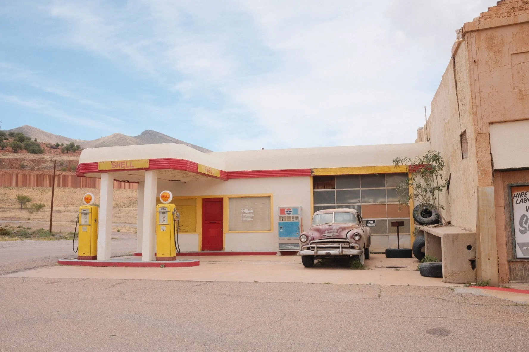 An old, deserted Shell gas station with yellow and red signage, two gas pumps, an abandoned vintage car, and tires stacked next to a weathered building in a barren, mountainous landscape.