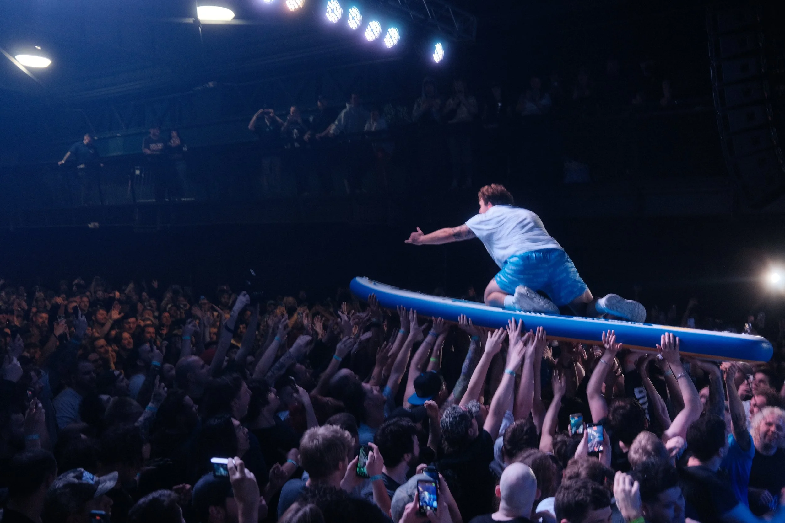 A performer is crouching on a surfboard stage, reaching towards a large crowd with outstretched hands, during a concert or live event. People in the audience are taking photos and videos, watching the performer on stage.