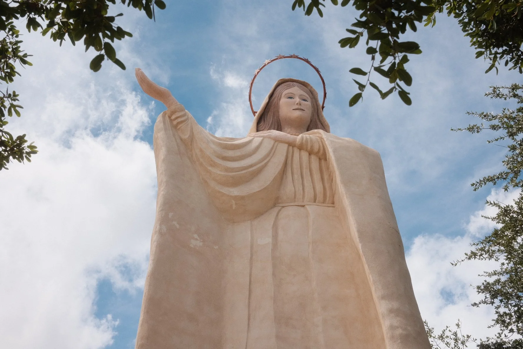 Statue of Jesus Christ with outstretched arms, overlooking the sky with clouds and framed by tree branches.