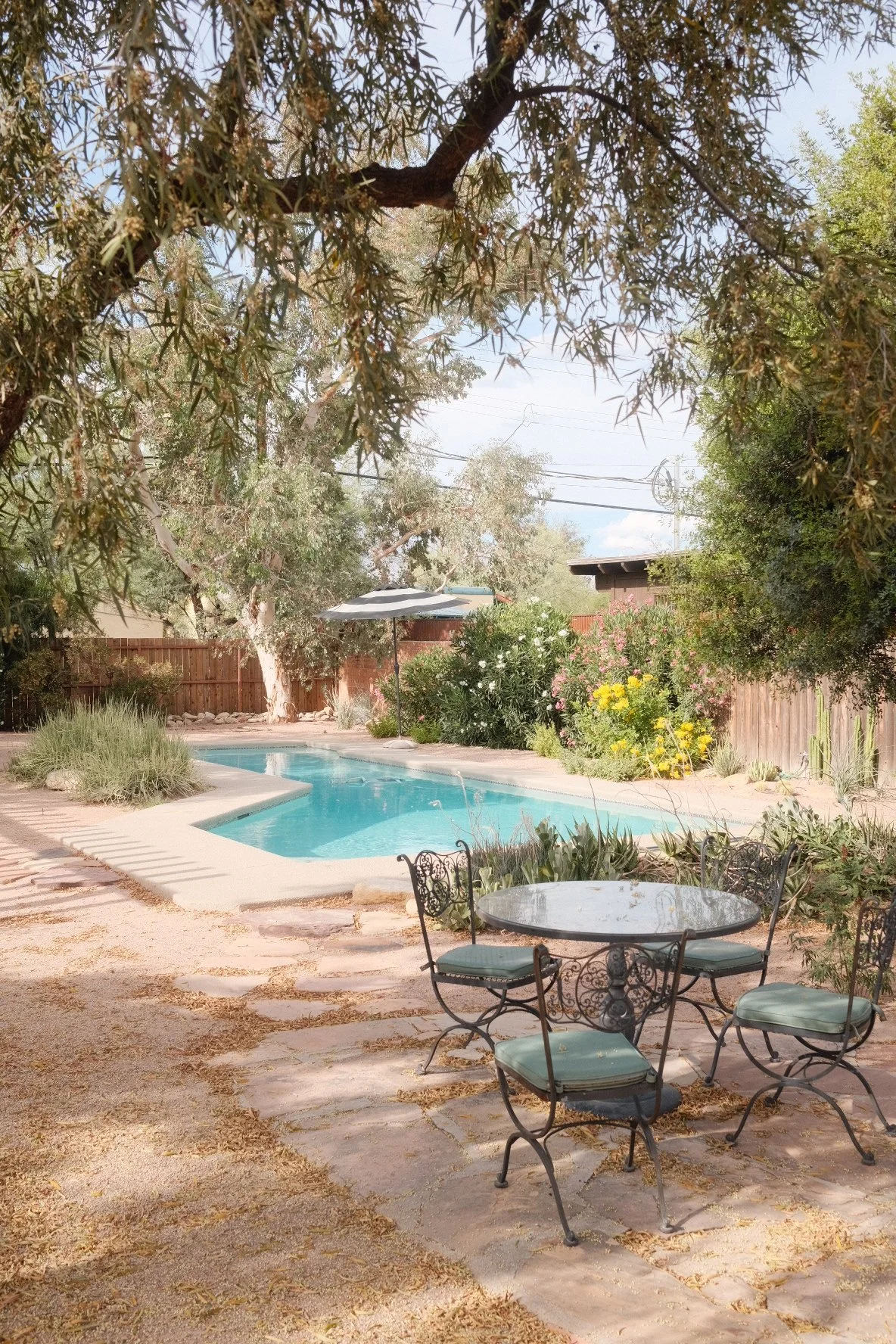 A backyard with a swimming pool, a patio table with four chairs, lush green trees and colorful flowers, and a wooden fence.