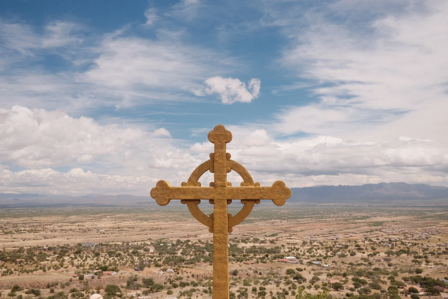 A metal cross-shaped icon with circular and decorative elements stands on a hilltop overlooking a desert landscape with sparse vegetation and distant mountains under a partly cloudy sky.