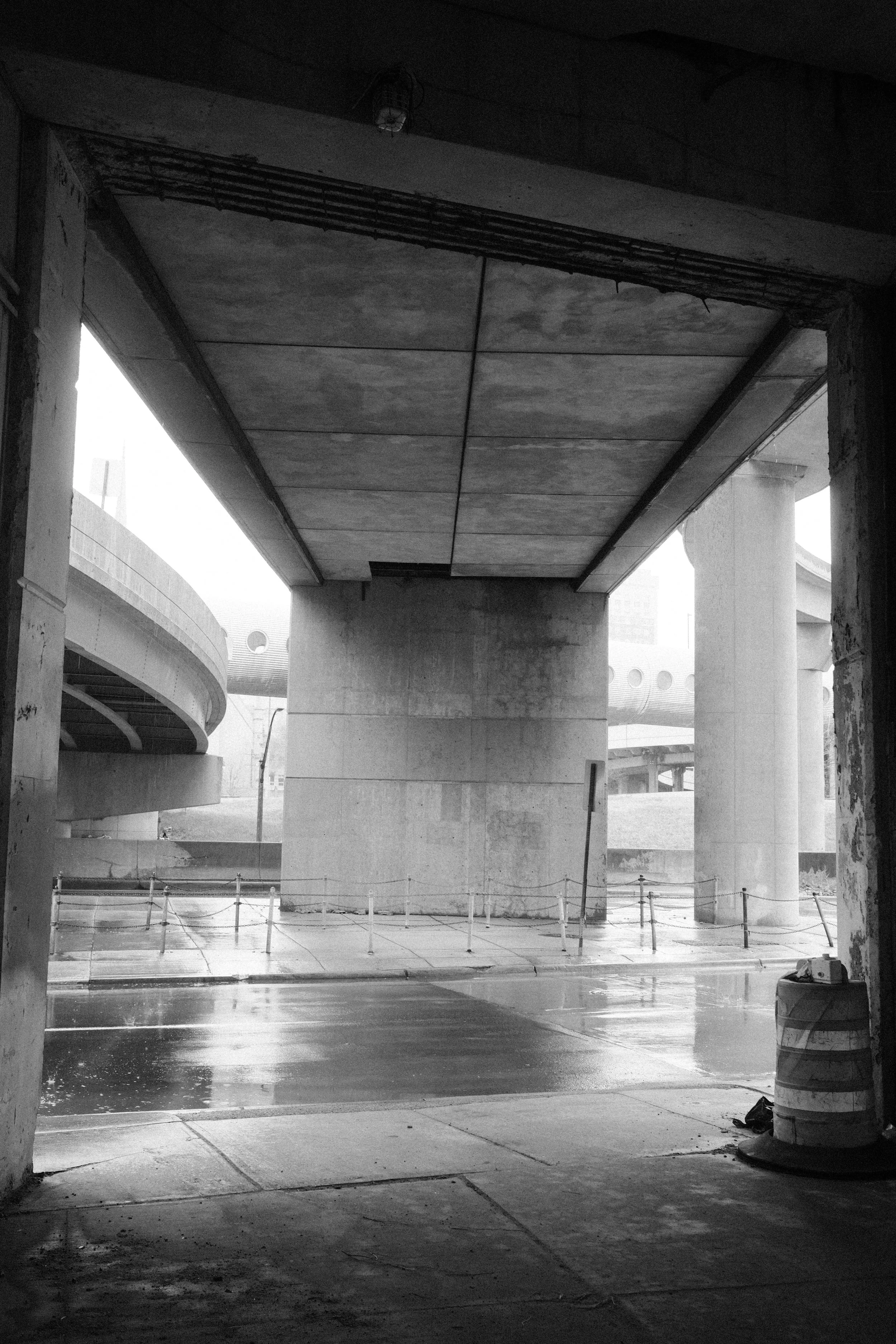Underpass construction with concrete pillars and a ceiling, some pipes and a light fixture, a wet floor, a chain-link barrier, and a hazy background with other structures.
