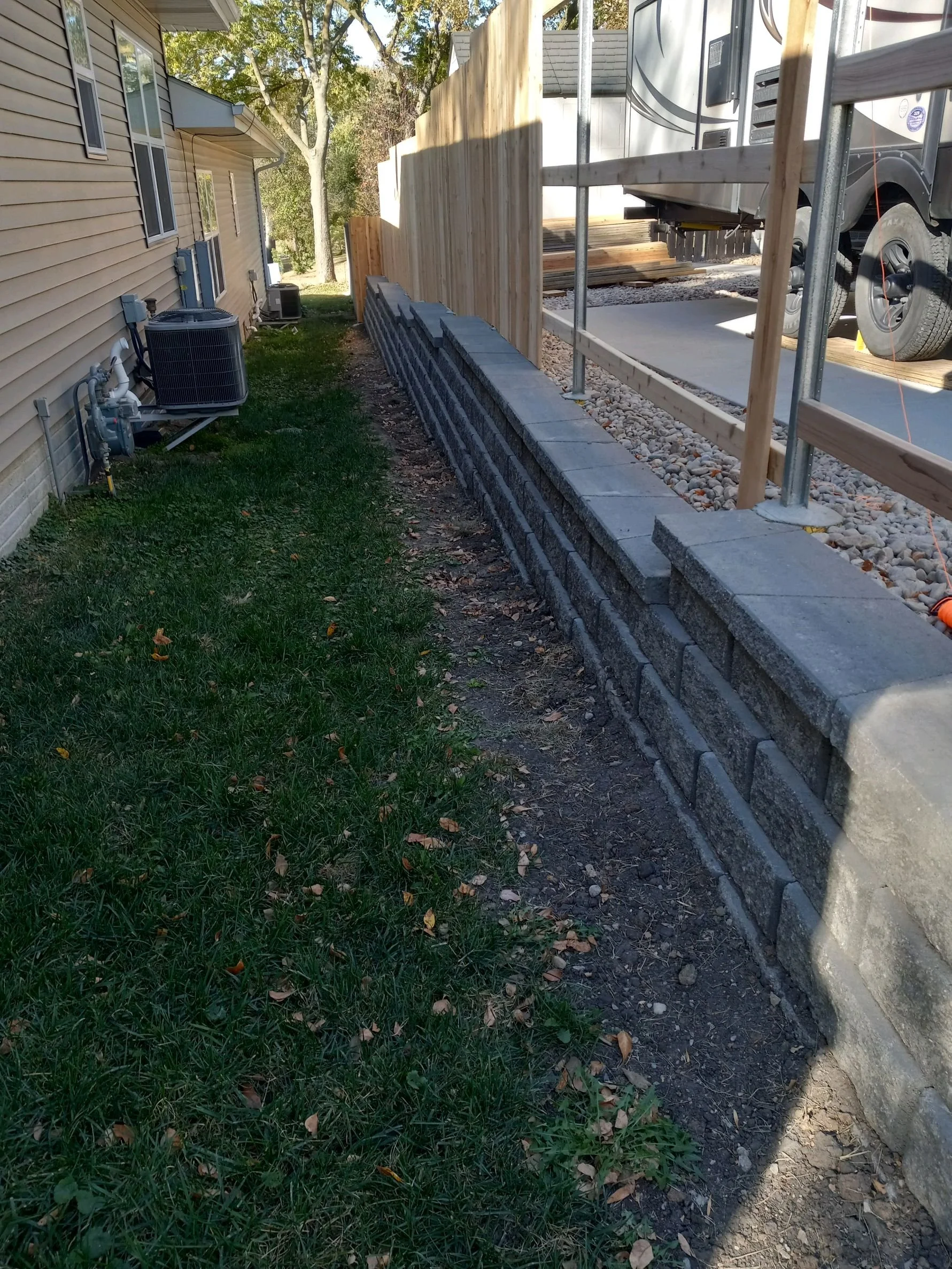 Side yard with green grass, a retaining wall, and a wooden fence next to a house and parked RV.