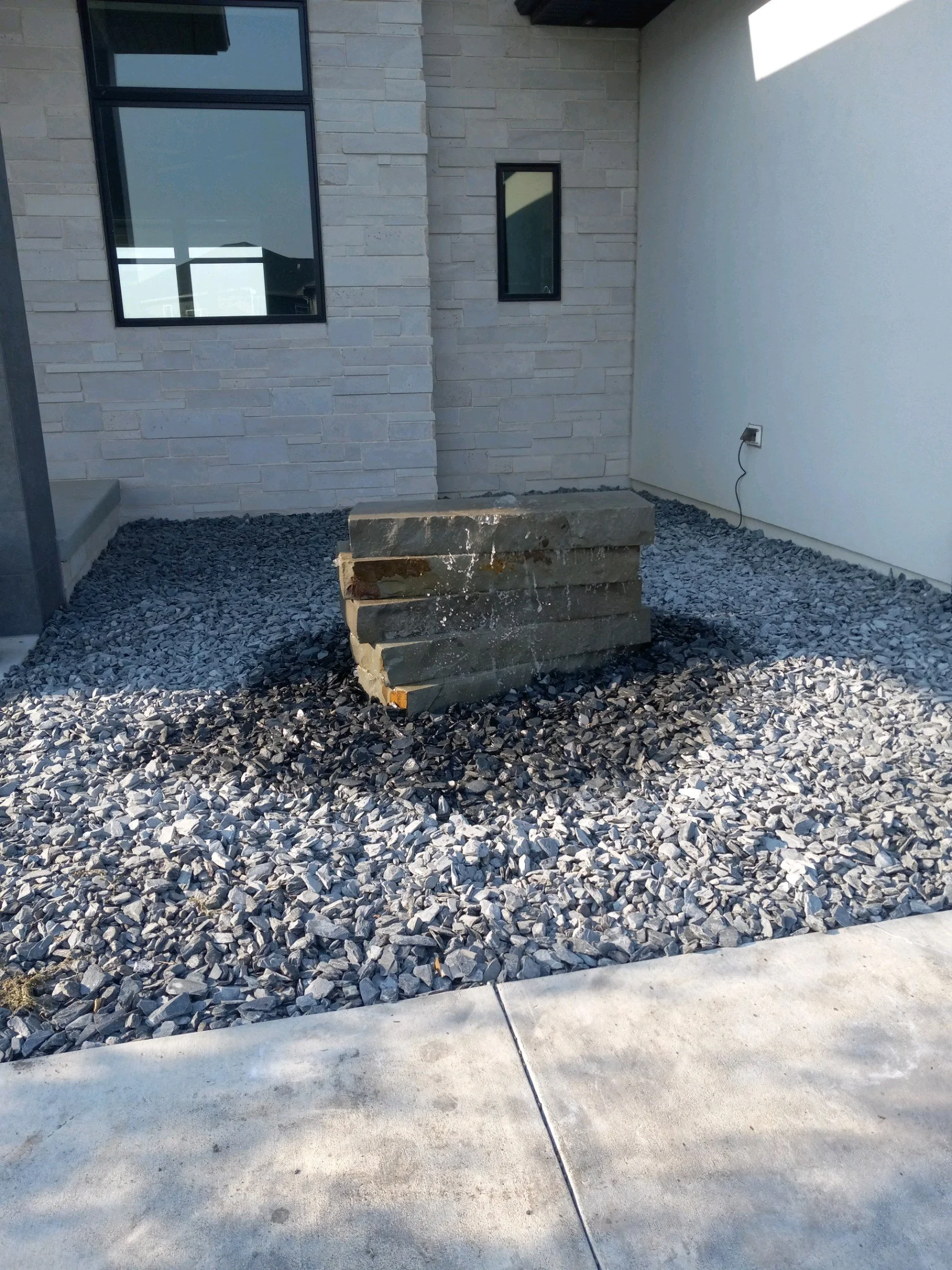 Outdoor stone fountain with water flowing, surrounded by gray gravel landscaping next to a modern building with stone cladding and windows.