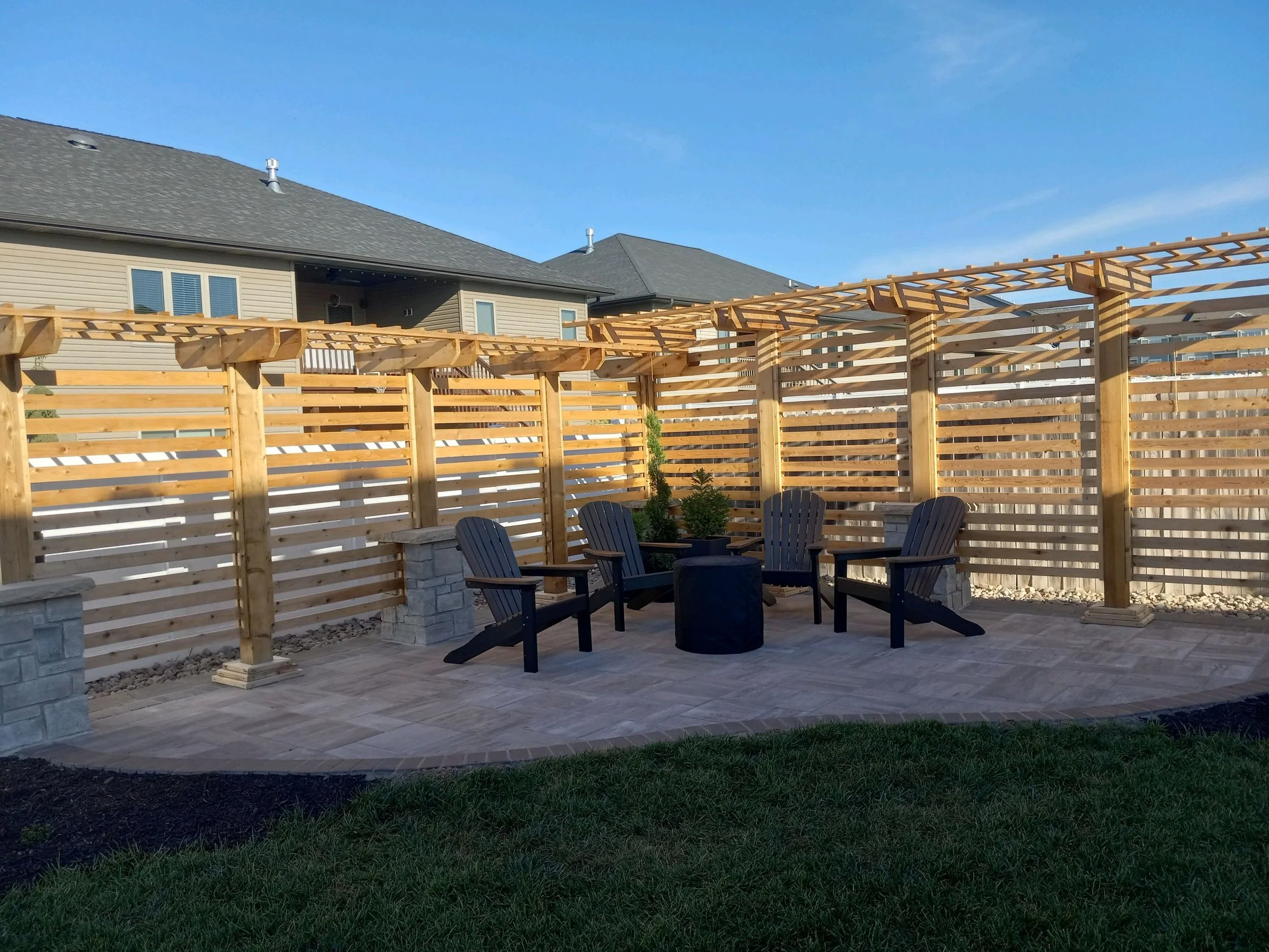 Backyard with a wooden pergola, four Adirondack chairs, a fire pit, and grass lawn.