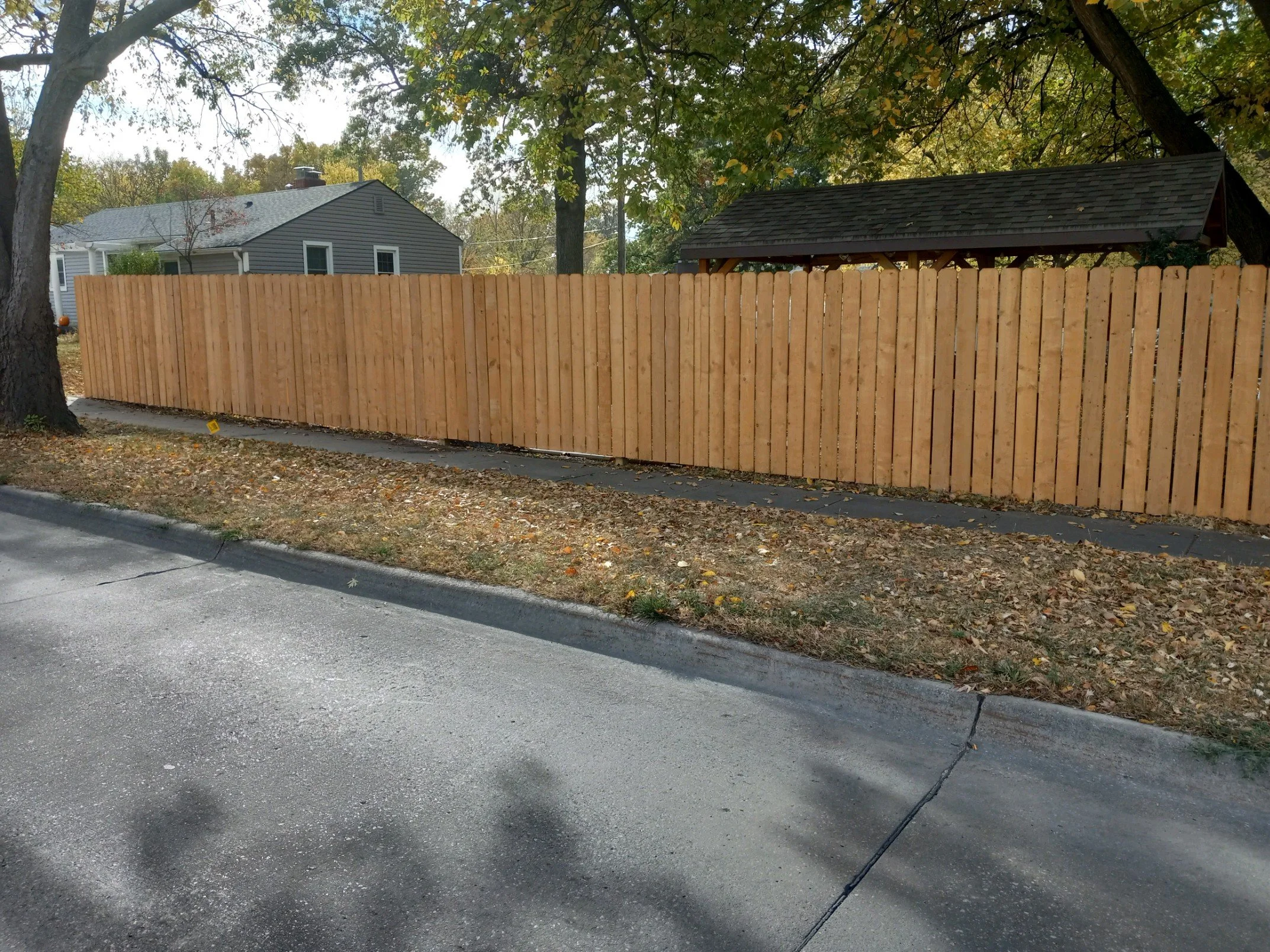 A wooden privacy fence along a sidewalk in a residential area, with a house and trees in the background.
