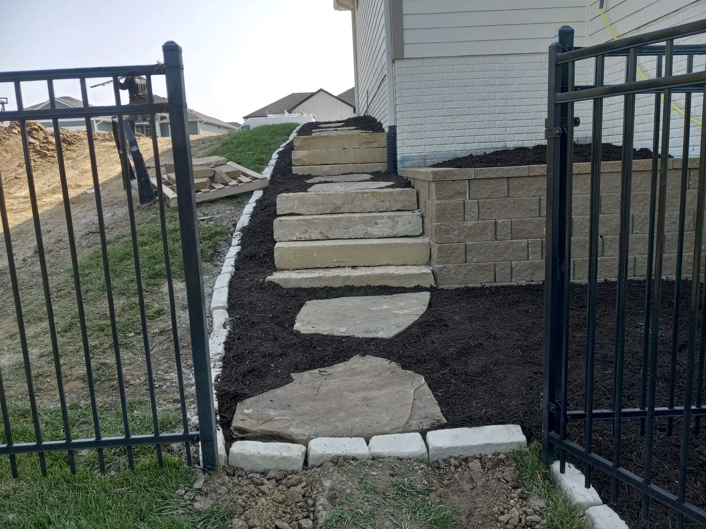 Stone pathway with stairs next to a house, bordered by a metal fence and landscaping mulch.