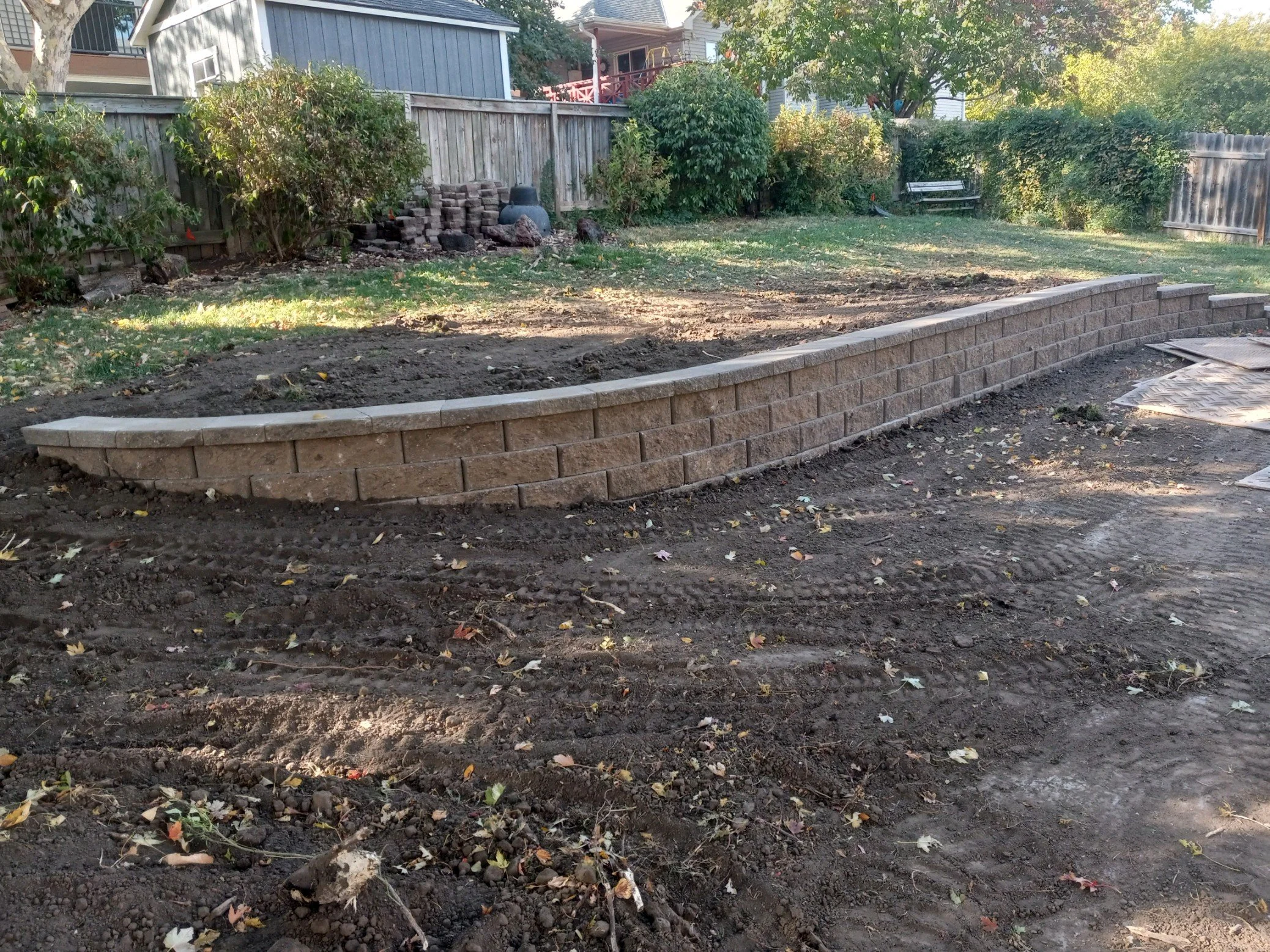 Brick retaining wall in a backyard under construction with dirt and scattered leaves.