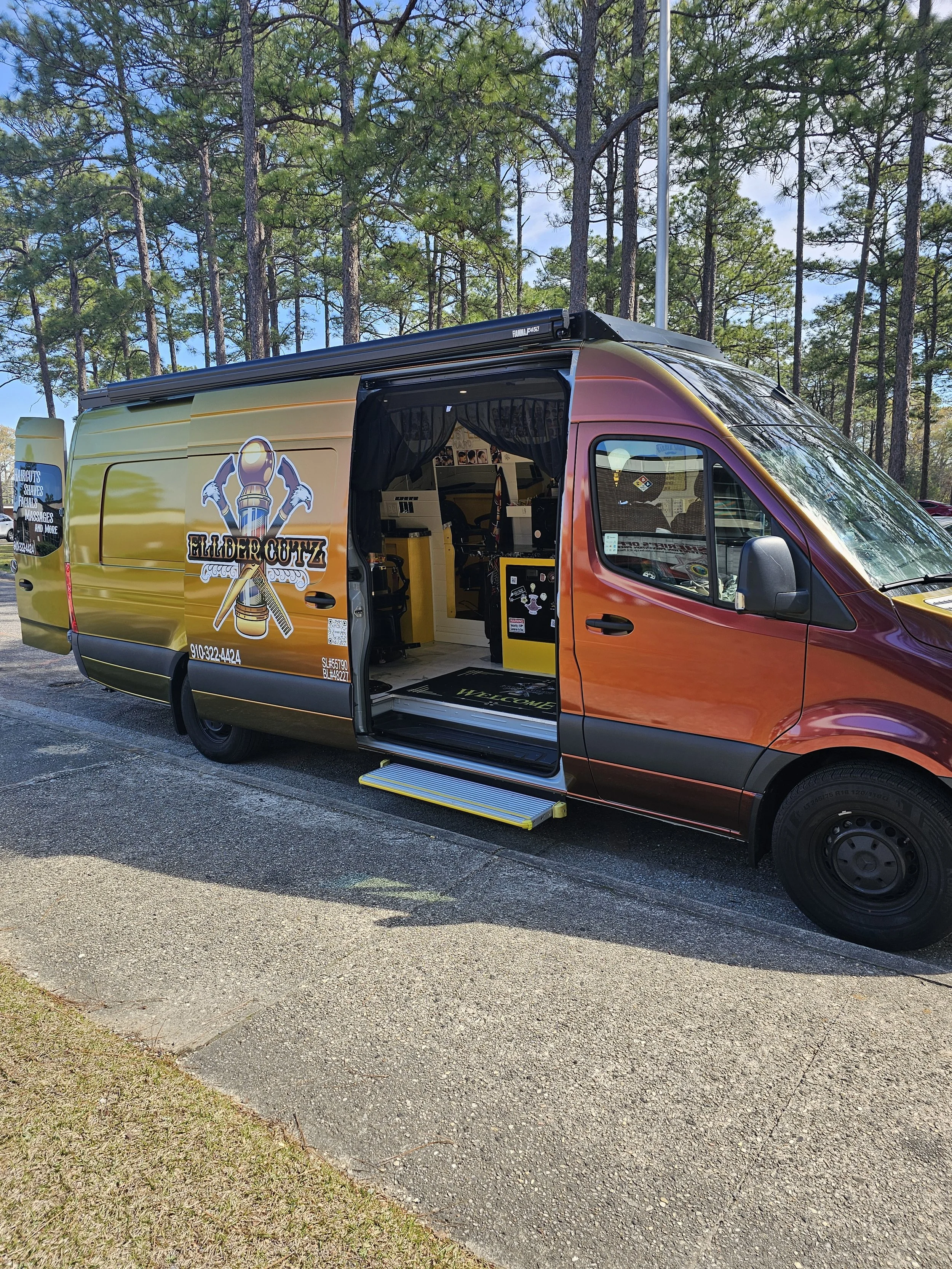 A yellow and orange mobile barber shop van parked in a forested area with tall pine trees in the background. The van's side door is open, revealing the interior.