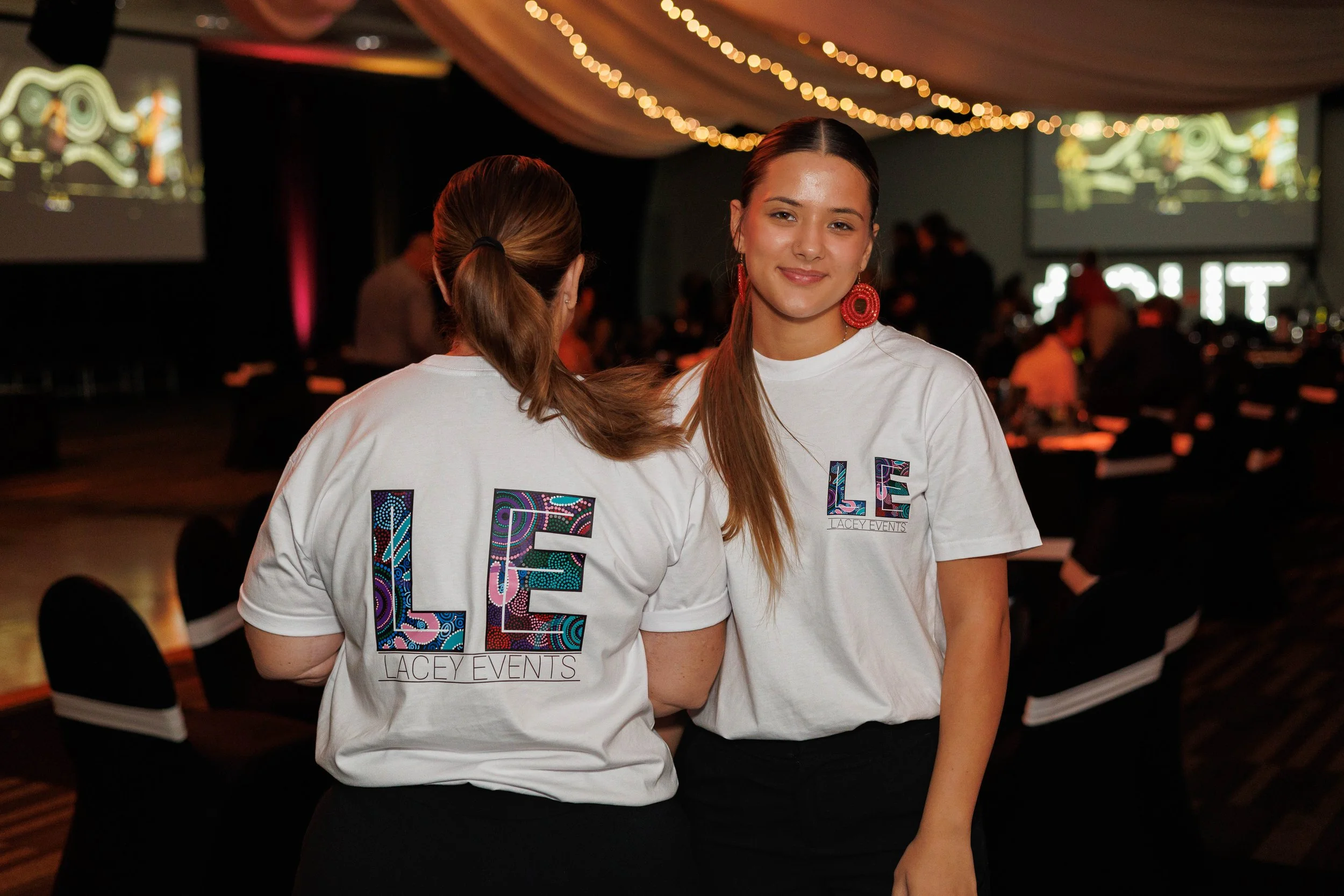 Two young women at an indoor event, wearing white t-shirts with colorful patterned letters that spell 'LOVE' and 'LACEY EVENTS.' One woman has long hair tied back and red earrings, smiling at the camera, while the other woman has her back to the camera with brown hair tied back. The background shows a stage with large screens, string lights, and other people in a dimly lit setting.