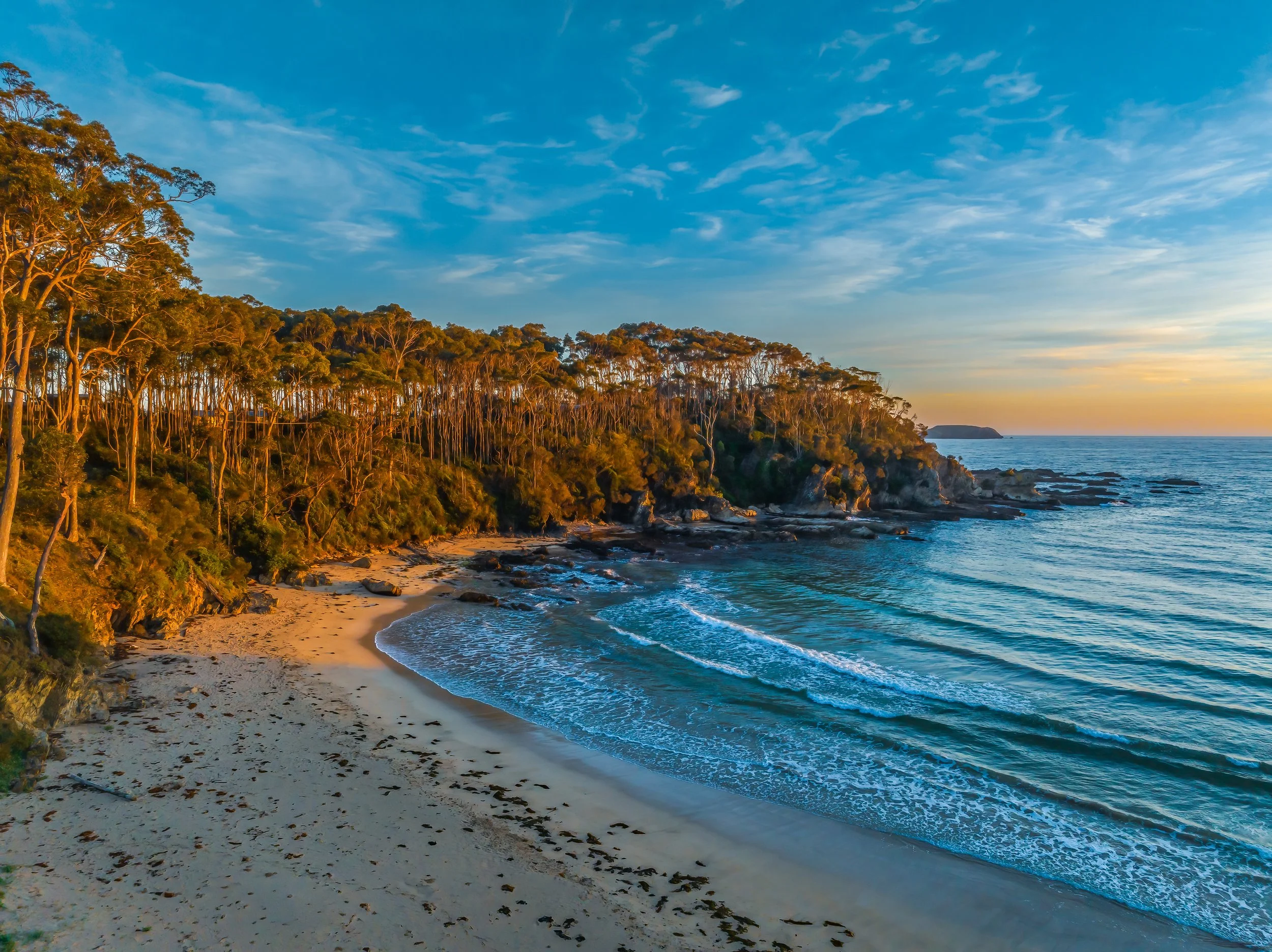 Sunset over a sandy beach with waves and a forested cliffside.