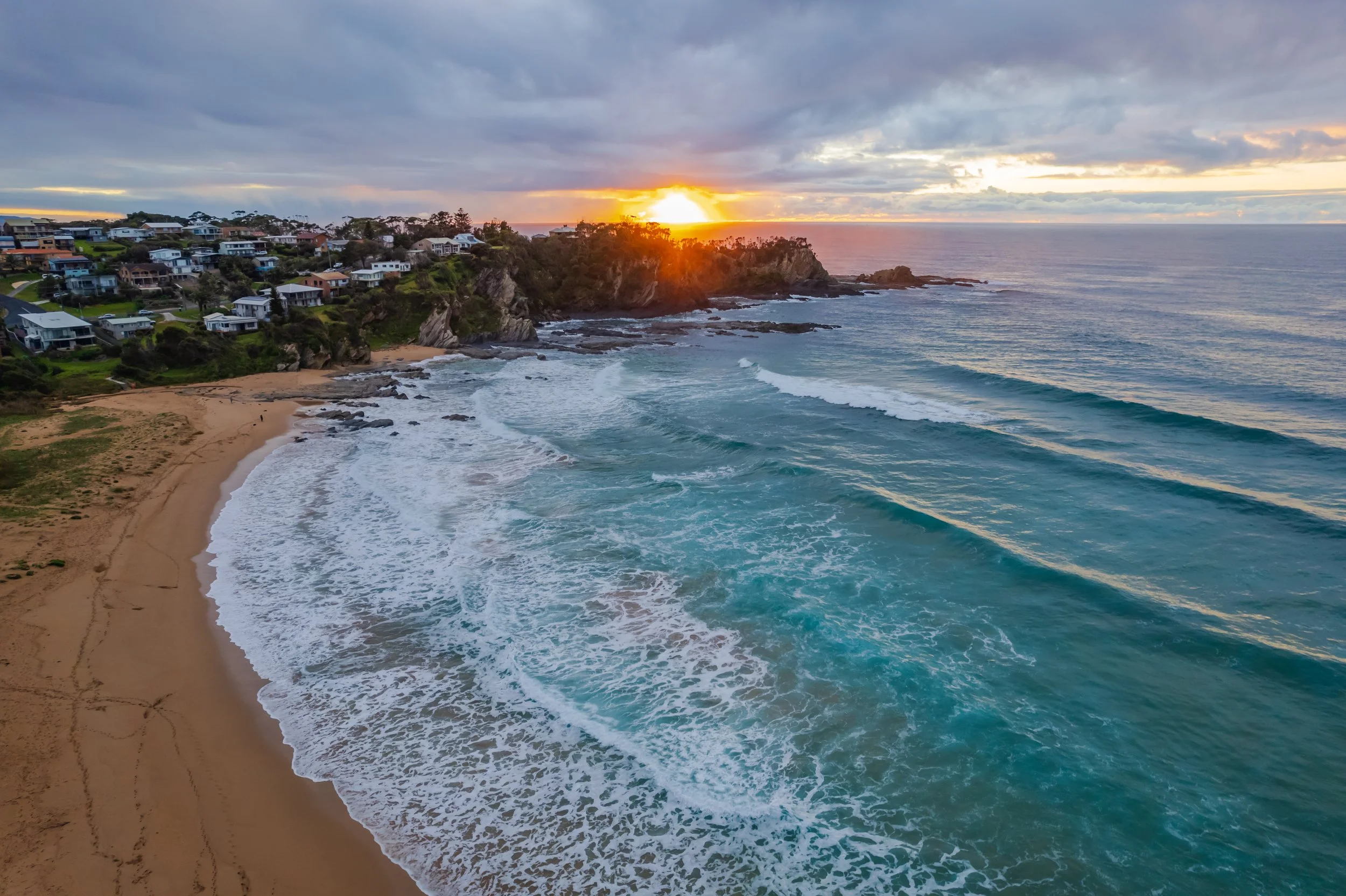 Sunset over a beach with waves crashing onto the sand, and houses on a hill overlooking the ocean.