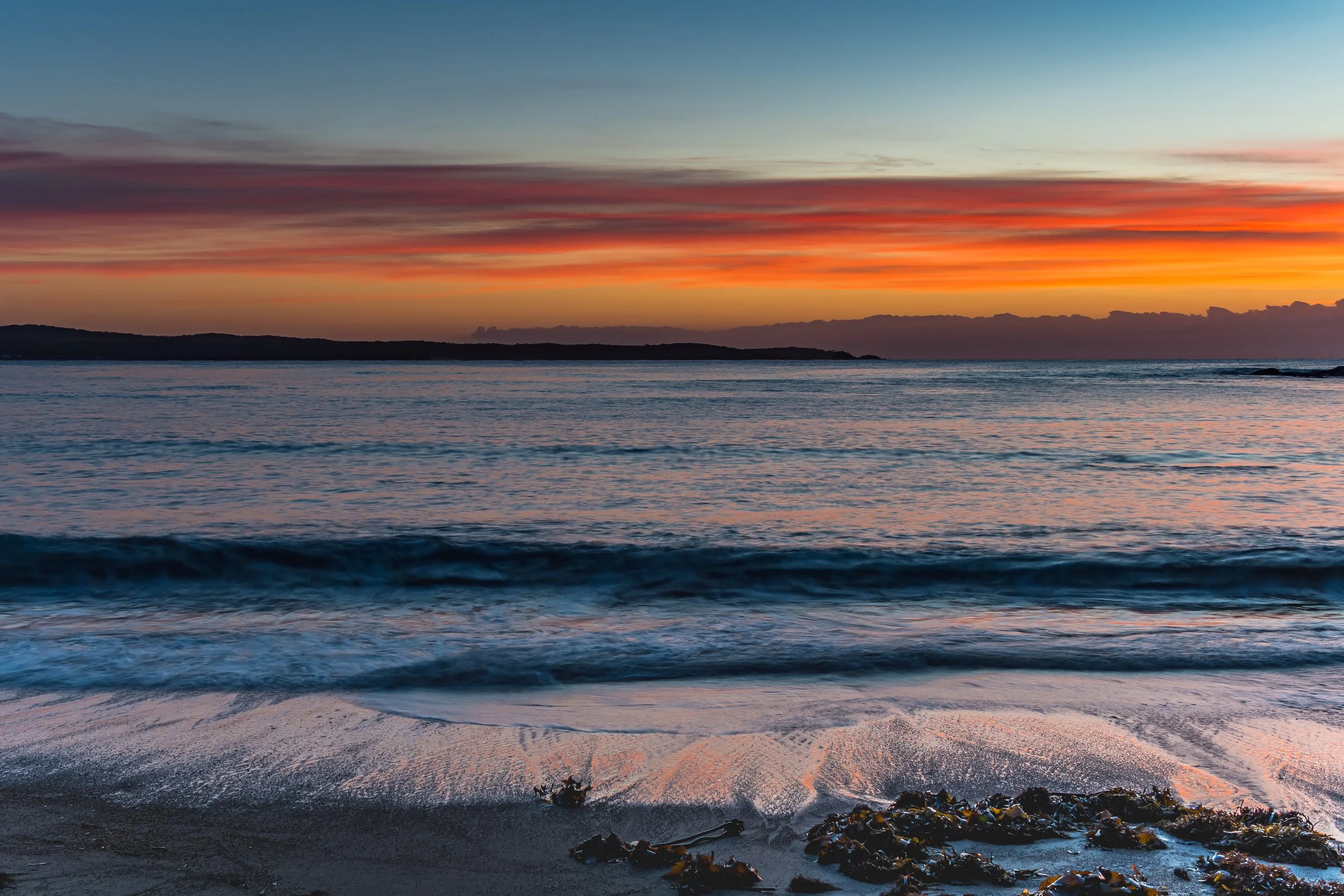 Sunset over the ocean with colorful sky, waves gently hitting the shore, and seaweed on the beach in the foreground.