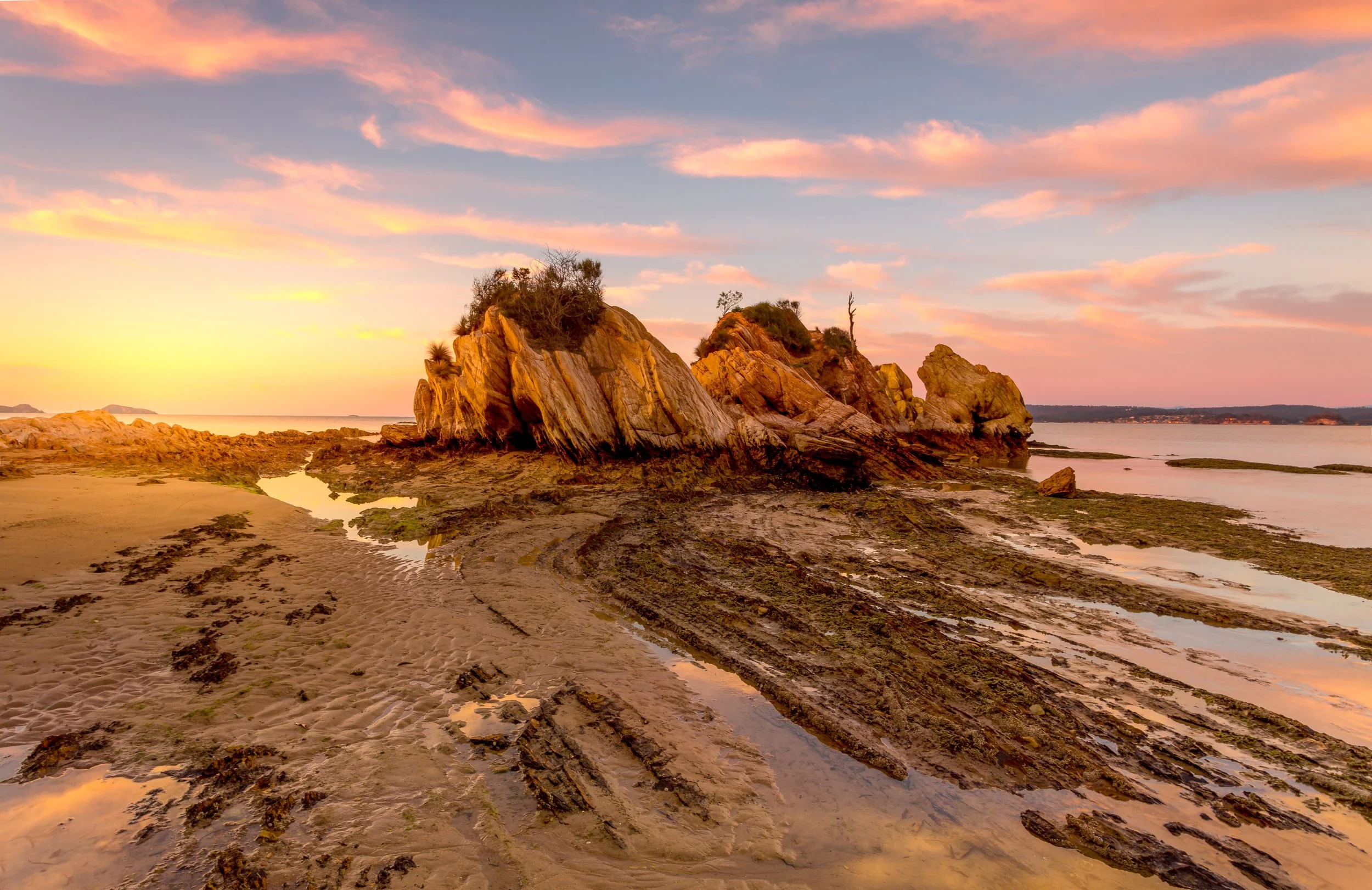Sunset over a rocky beach with tide pools and a large rock formation with sparse vegetation.