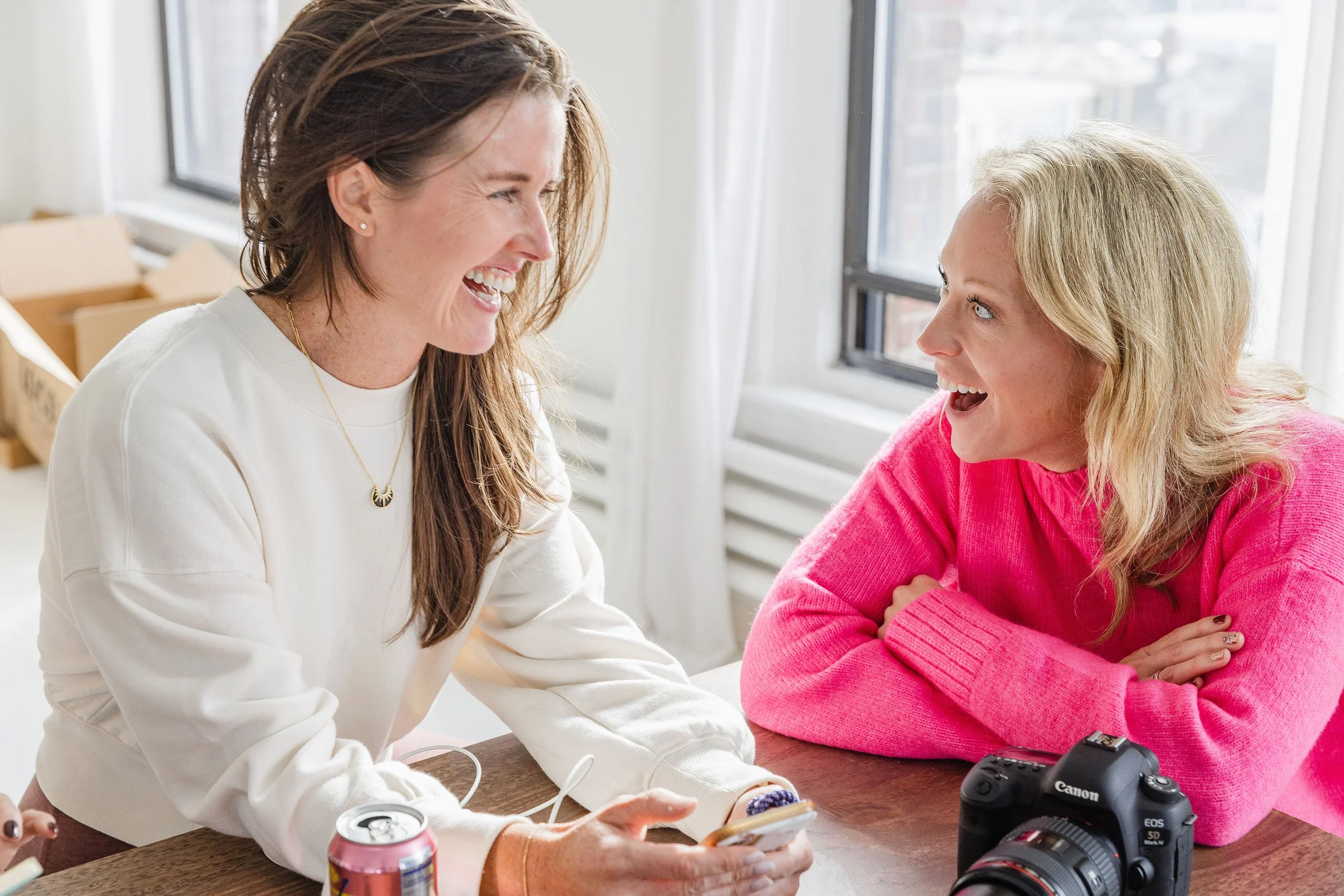 Two women smiling and talking across a table in a bright room with windows, one wearing a white sweatshirt and the other in a pink sweater, with a camera and soda cans on the table.