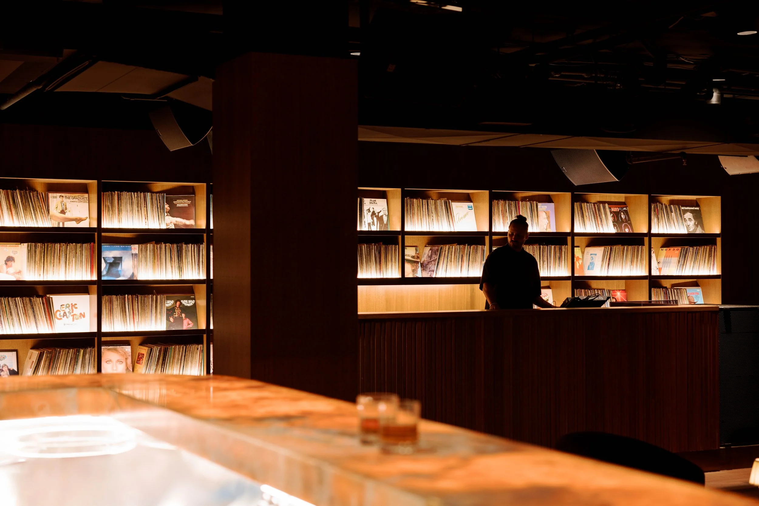 A dimly lit room with wooden shelves filled with vinyl records, a person standing behind a wooden bar or counter, and a table with two glasses in the foreground.