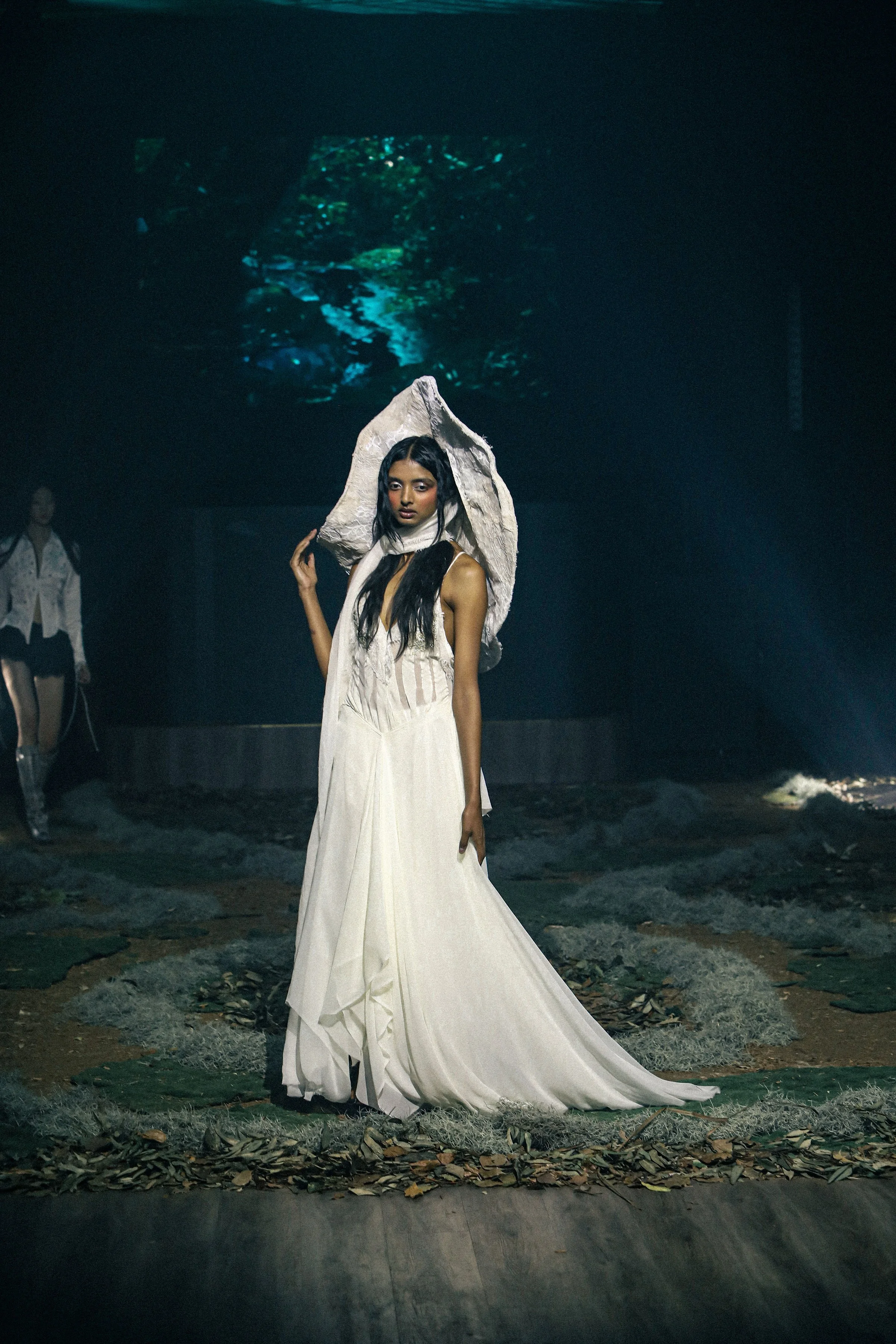 A model walking on a runway wearing a long, flowing white dress and a large stone-like headpiece, with dark surroundings and a nature-themed backdrop.