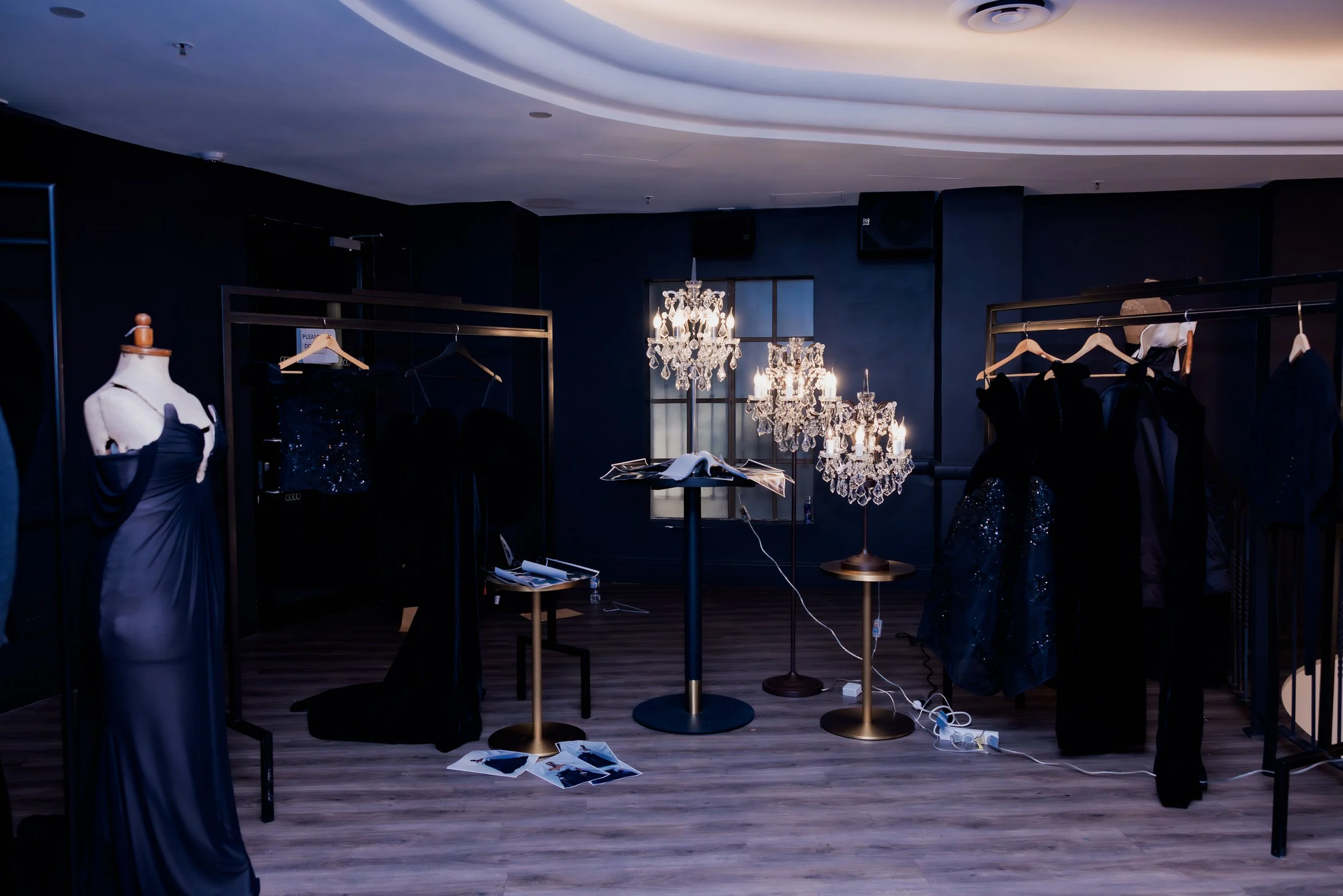 Dimly lit room with black walls displaying elegant black evening dresses and outfits on mannequins and racks, illuminated by ornate chandeliers and standing lamps.