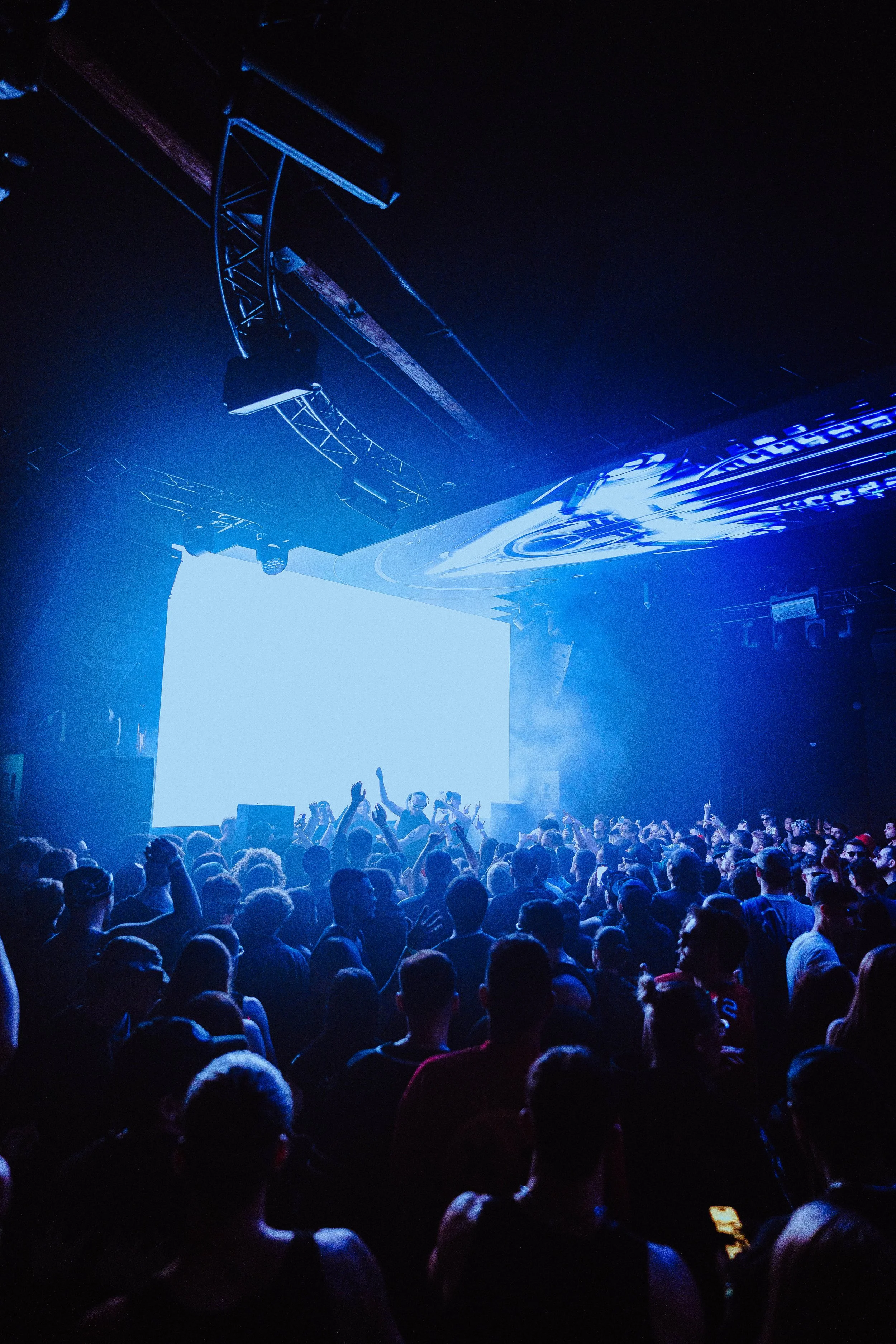Crowd of people at a concert or music event with bright blue stage lights and large screen in the background.