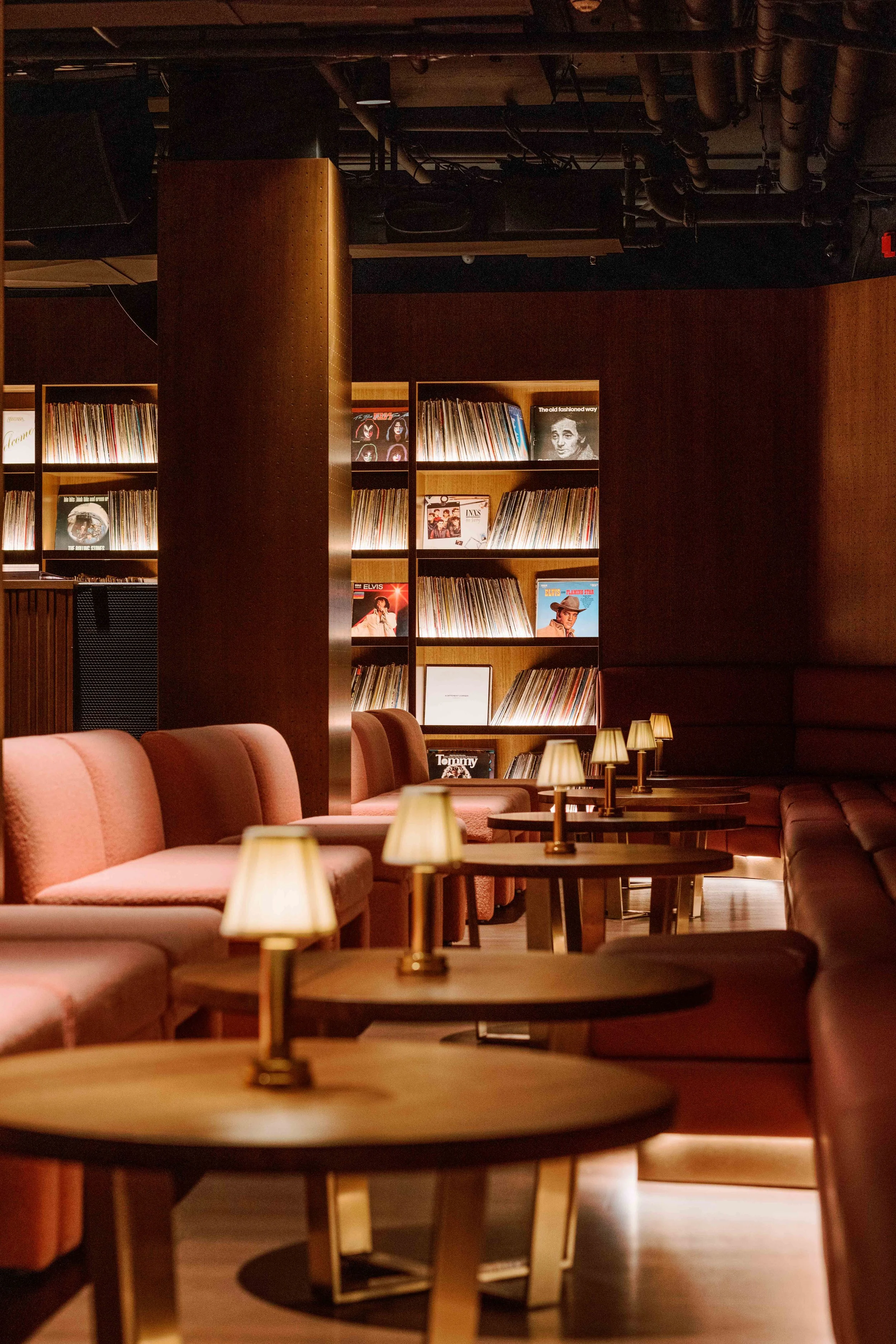 A cozy lounge area with pink cushioned chairs, small tables with lamps, and a background of wooden shelves filled with vinyl records.
