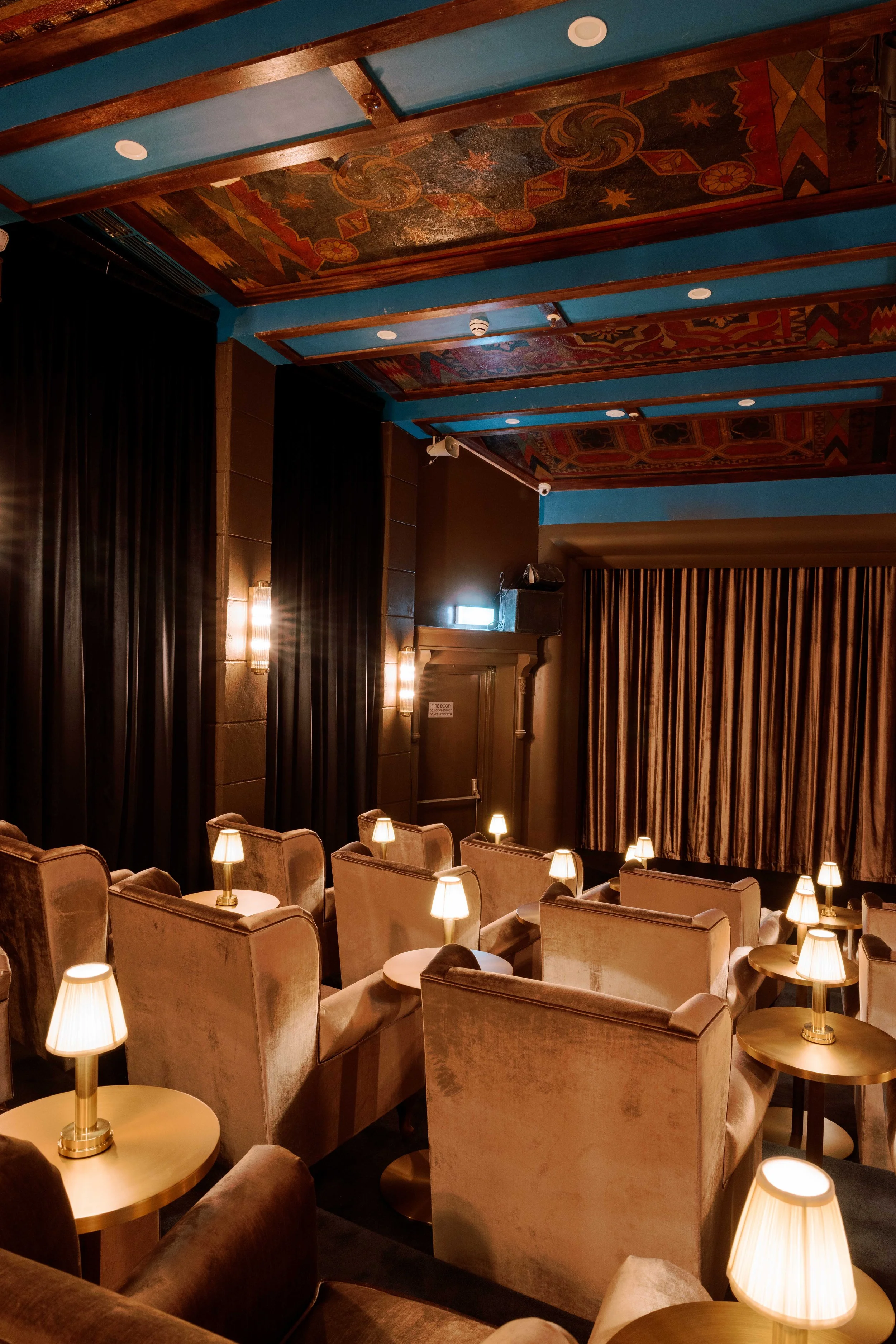 A cozy movie theater with plush beige armchairs arranged in rows, each equipped with small side tables with lamps. There are tall black curtains on the walls, a stage with closed curtains, and a decorative painted ceiling with modern geometric art and warm lighting.
