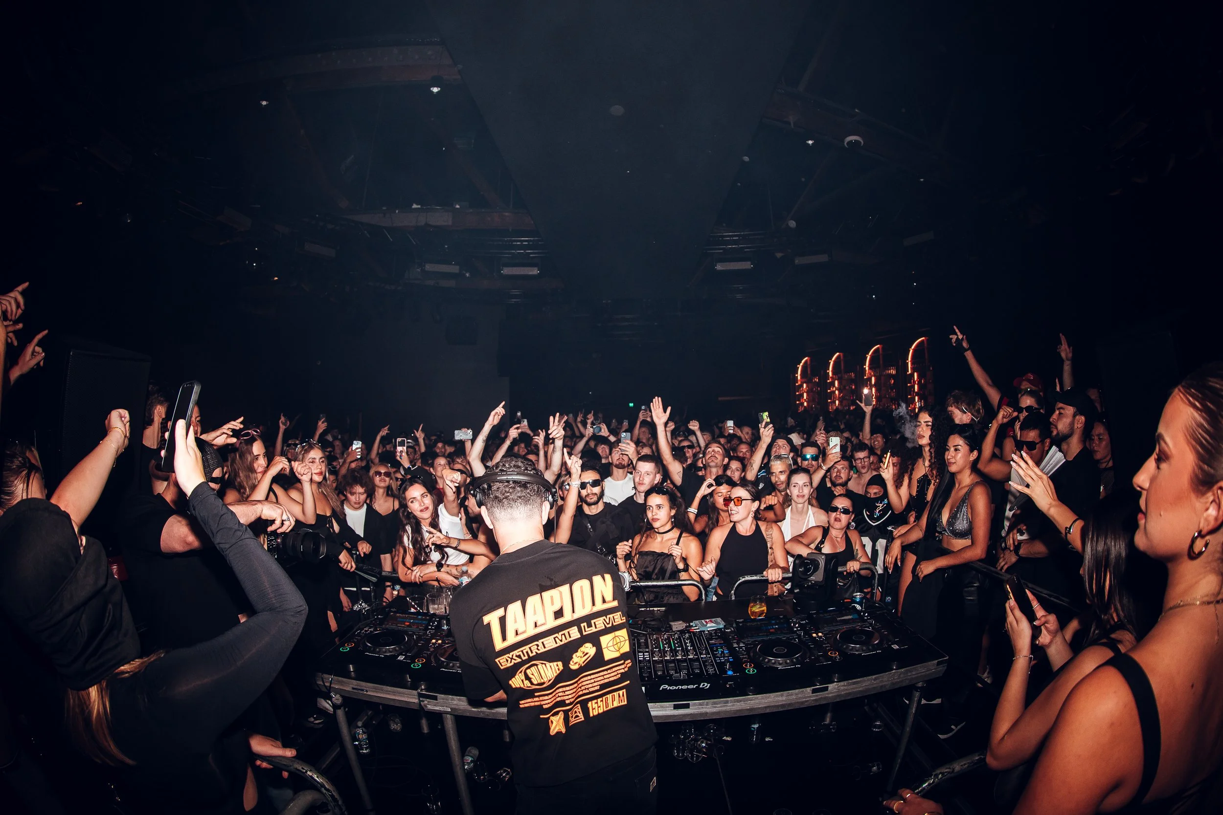 Crowd of people at a concert dancing and taking photos with a DJ performing in the foreground.