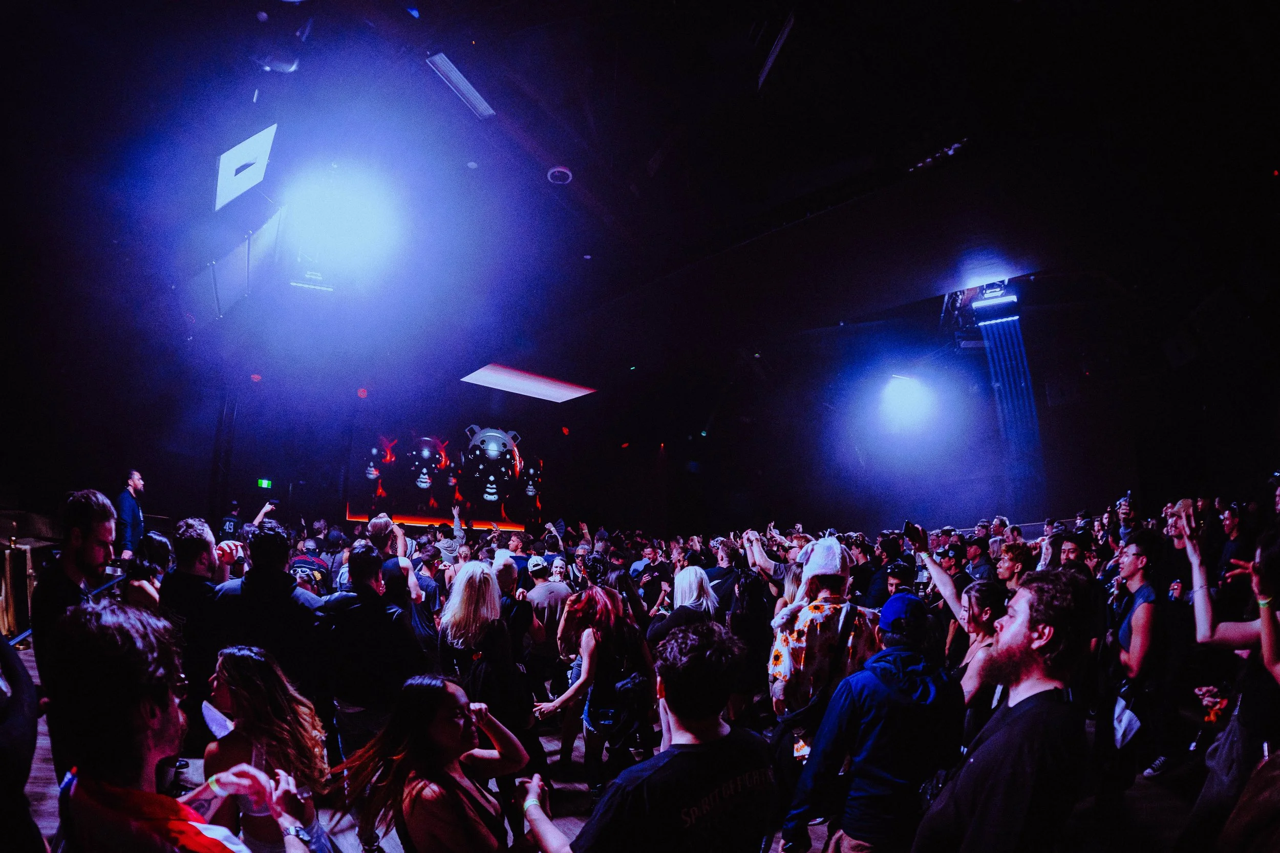 Crowd attending a concert or DJ event at a nightclub with blue and purple lighting.